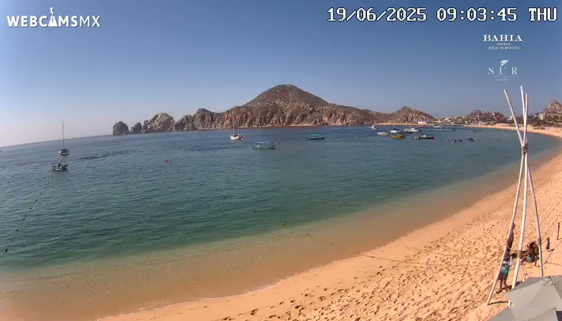 A sandy beach curves gently around a calm, clear bay with several boats, under a clear blue sky.