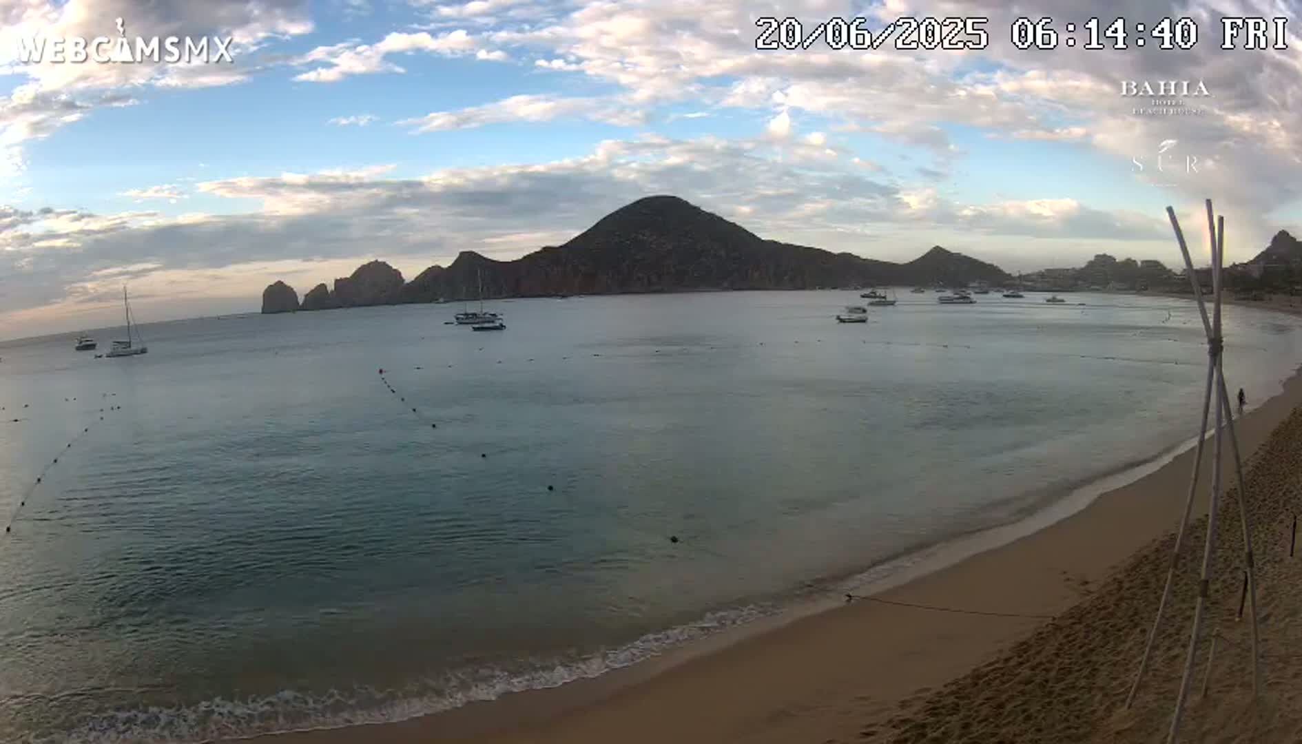 A sandy beach with calm, shallow water, several boats in the bay, and a large landmass in the background under a partly cloudy sky.