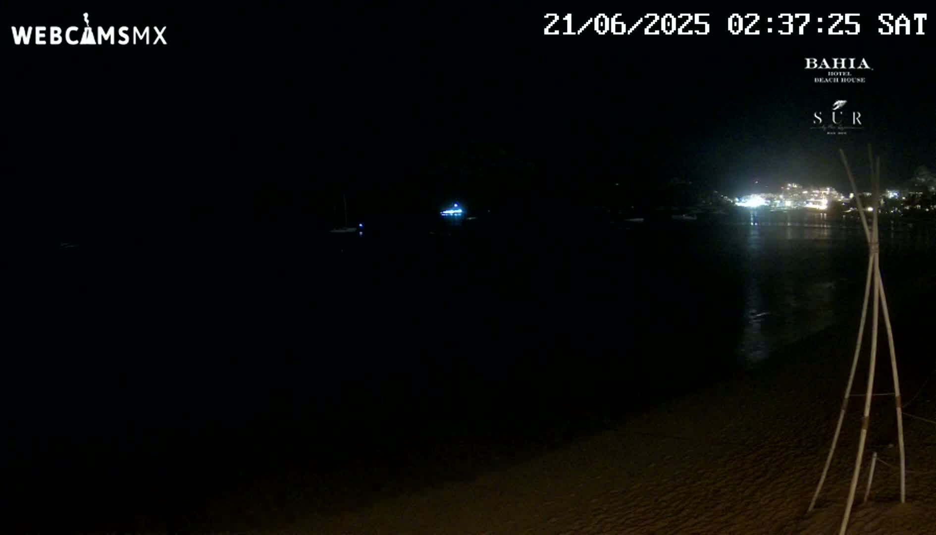 A dark nighttime beach scene shows a calm body of water reflecting distant lights, with a sandy shore in the foreground and a tripod-like structure to the lower right.