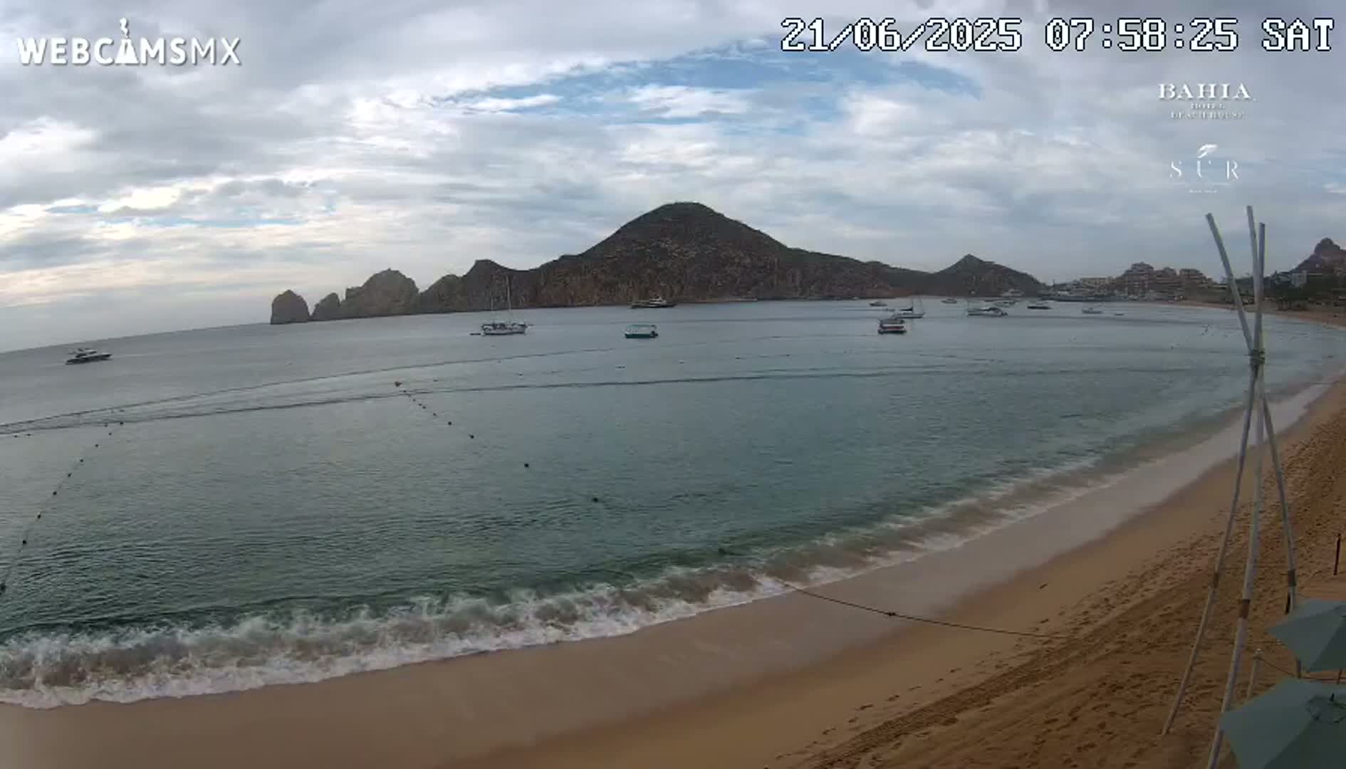 A sandy beach with calm, gentle waves laps the shore under an overcast sky, showcasing a bay with several boats and a mountainous backdrop.