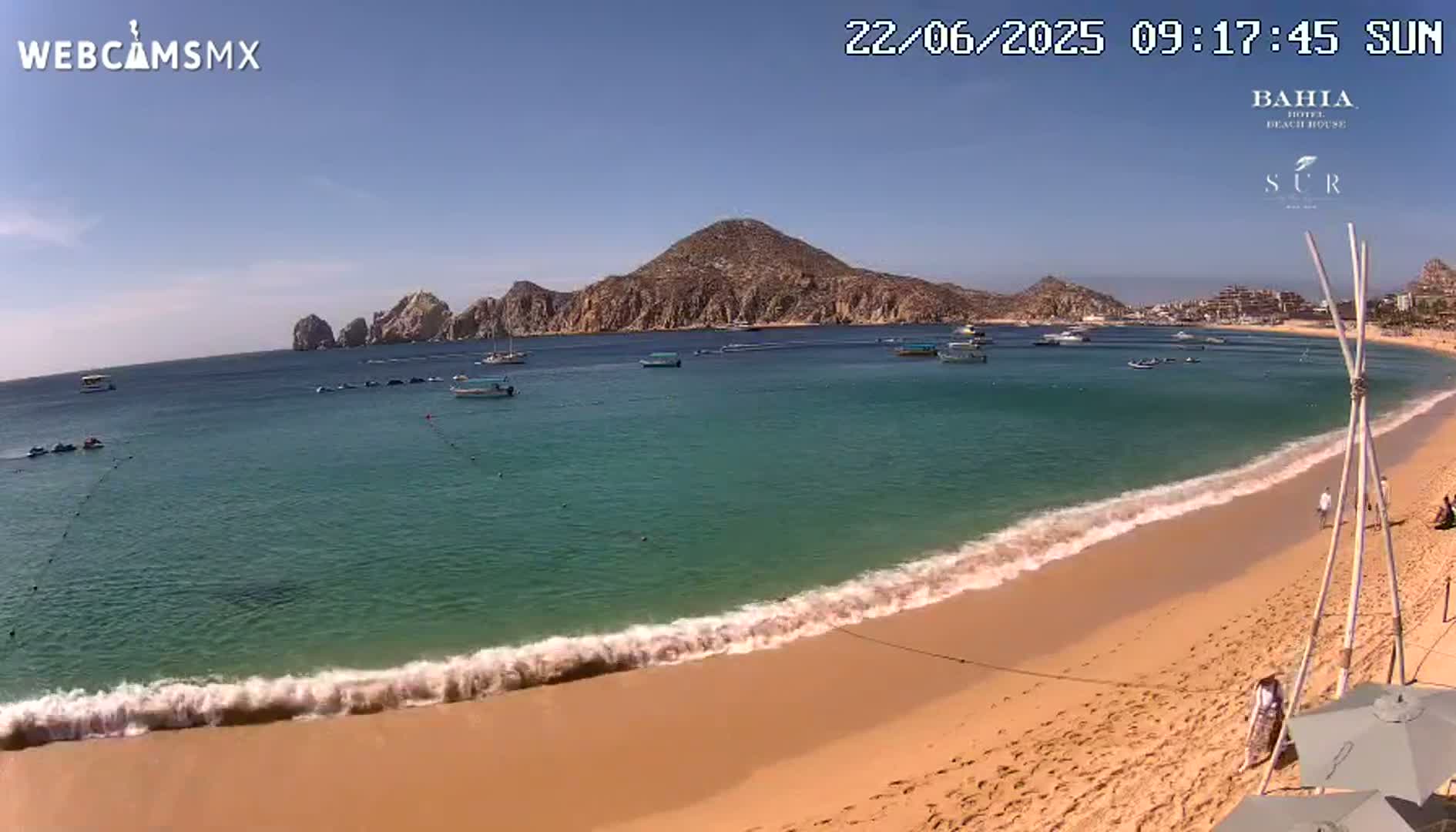 A sandy beach with calm, turquoise water laps gently at the shore under a clear, sunny sky, with boats and a rocky landmass visible in the distance.