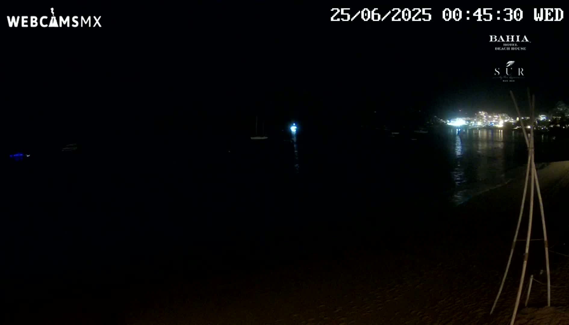 A dark nighttime beach scene shows a calm body of water reflecting the lights of a distant town, with a few boats visible on the water.