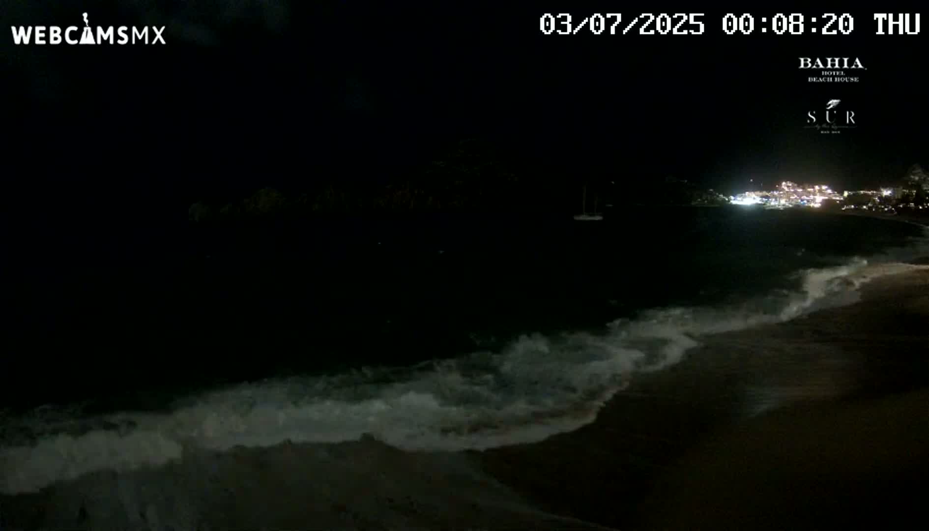 A nighttime beach scene shows dark waves lapping onto a shore with lights visible across the water in the distance.