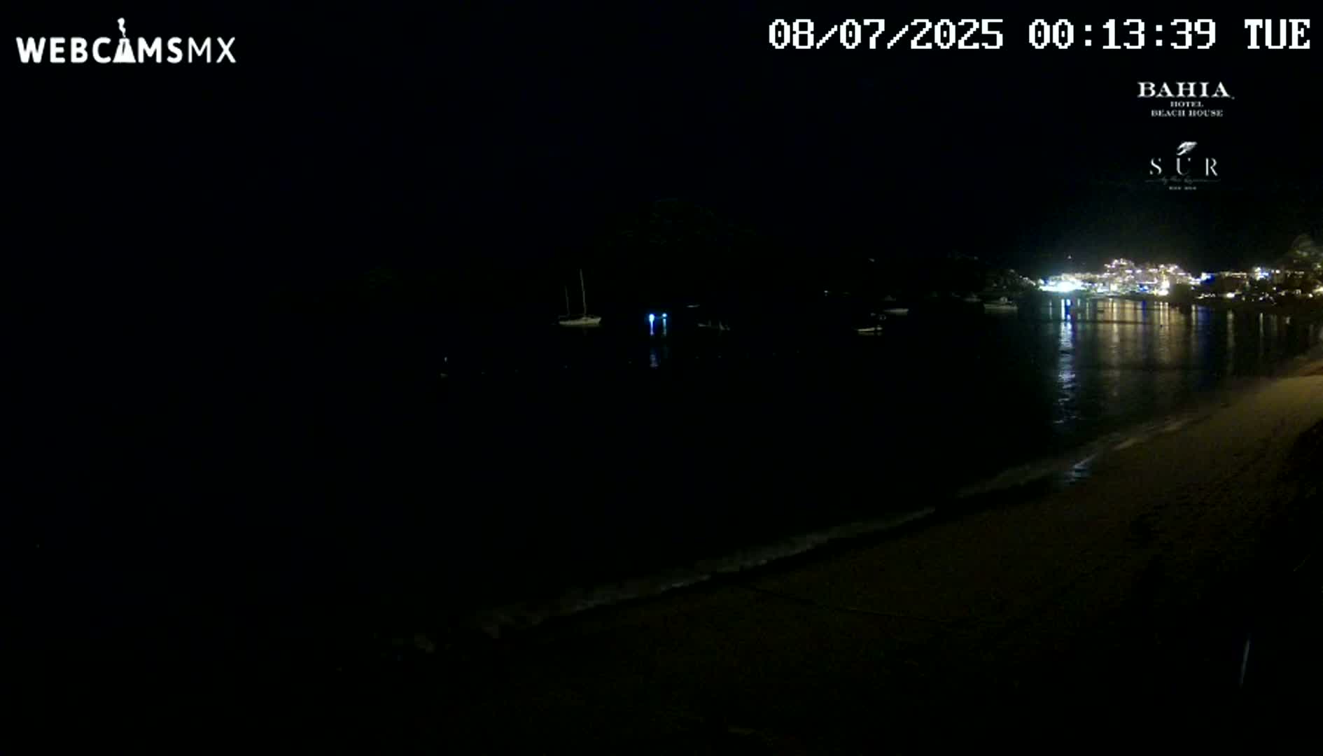A dark beach at night is shown, with calm water reflecting the lights of buildings across the bay.