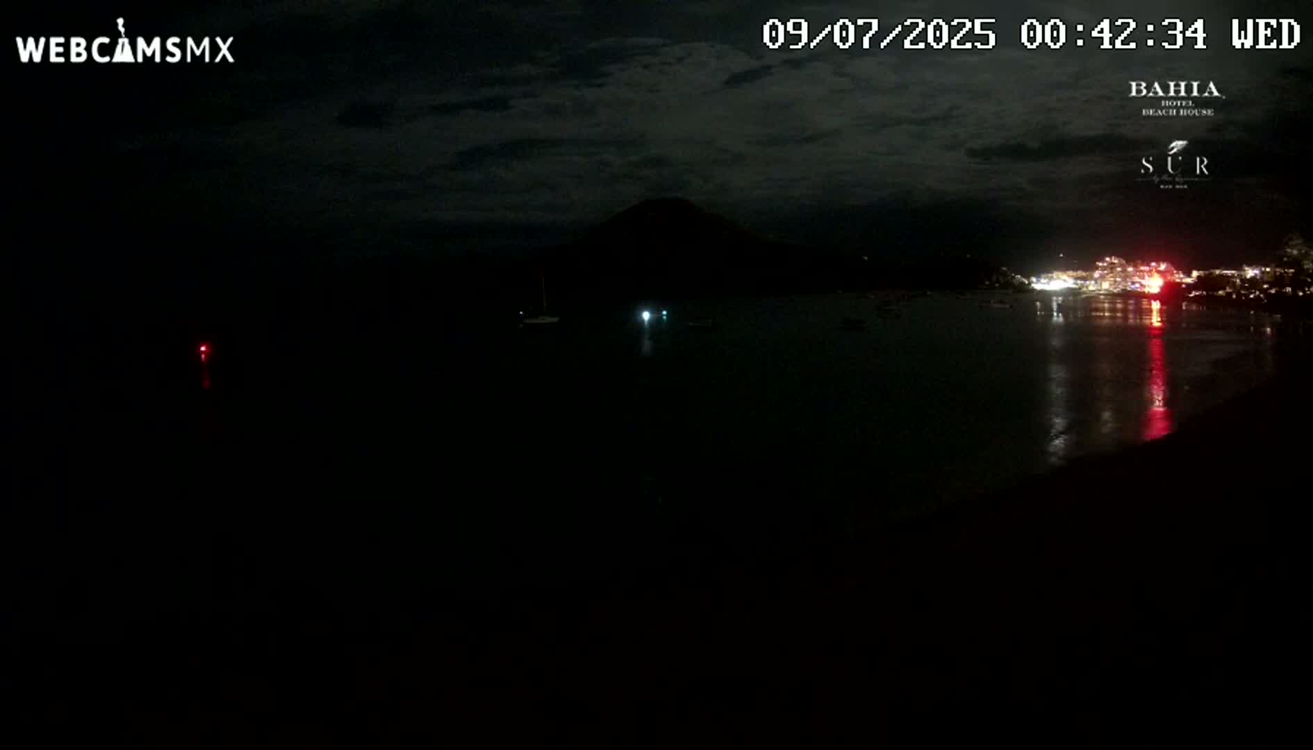 A dark nighttime scene shows a calm body of water reflecting the lights of a distant town under a cloudy sky, with a dark mountain silhouetted in the background.