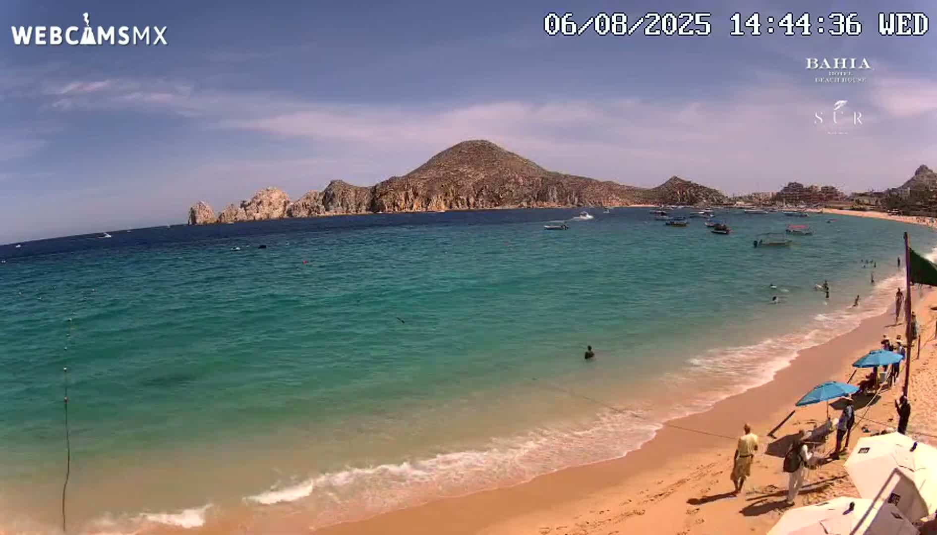 A sandy beach with calm, turquoise water, several people swimming and relaxing, and boats in a bay under a sunny sky.