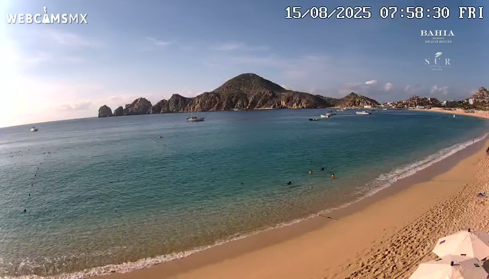 A sandy beach with calm, turquoise water, several people swimming and relaxing, and boats in a bay under a sunny sky.