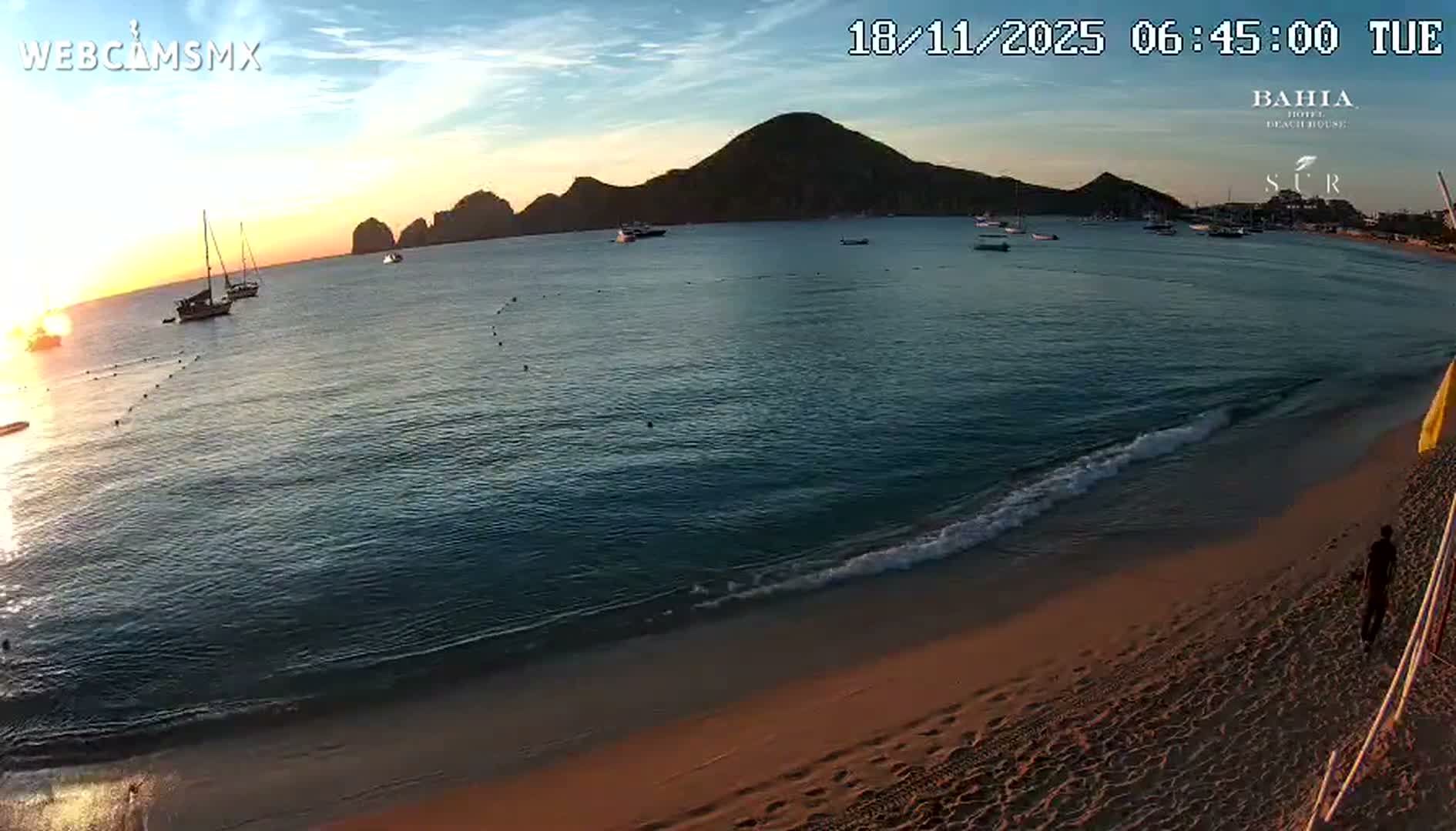 A sandy beach with calm, turquoise water, several people swimming and relaxing, and boats in a bay under a sunny sky.