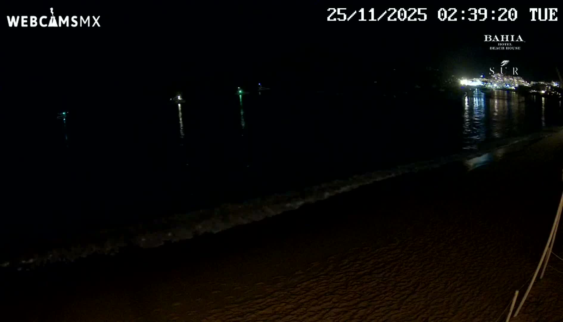 A nighttime view of a beach features gentle waves washing ashore, with distant city lights reflecting on the water and a few illuminated boats visible in the bay under clear skies.