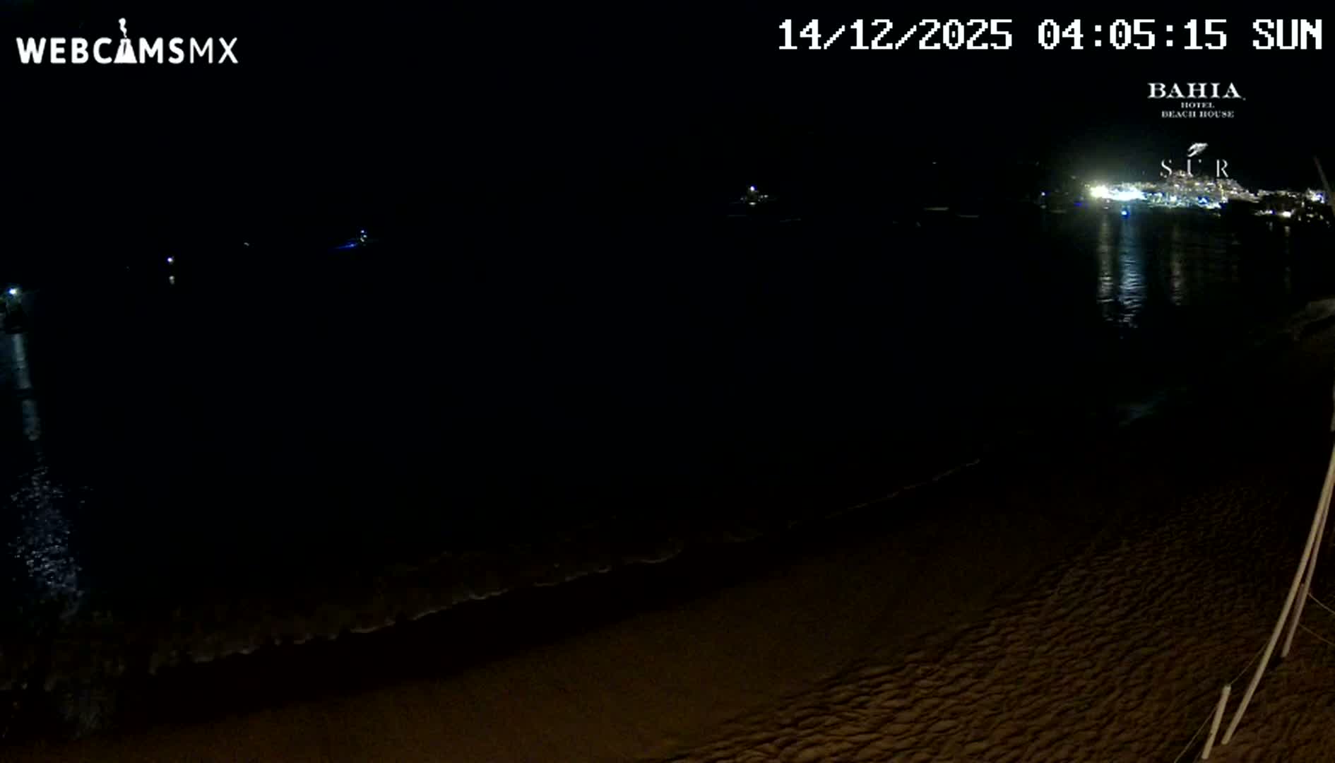 A nighttime view of a beach features gentle waves washing ashore, with distant city lights reflecting on the water and a few illuminated boats visible in the bay under clear skies.