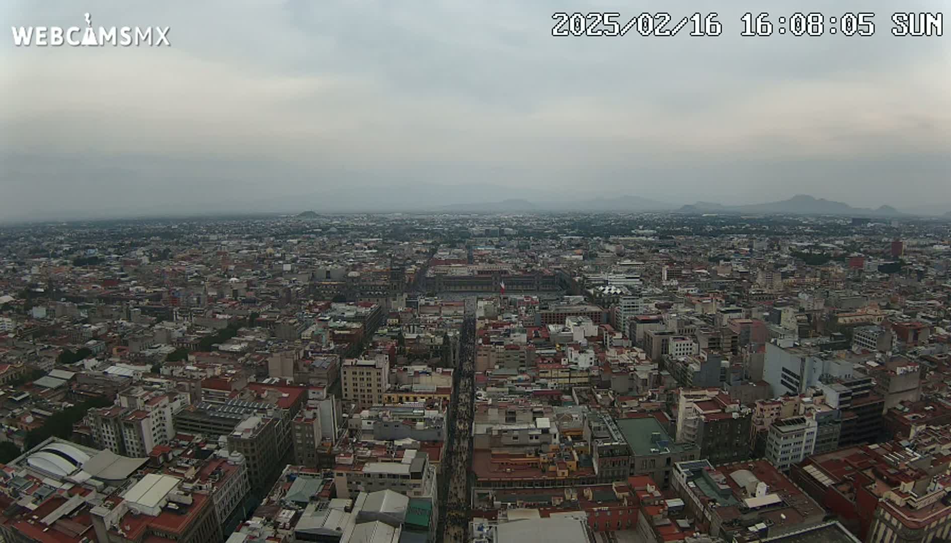 A high-angle, long shot on a cloudy day shows a sprawling city with a large plaza in the center, mountains visible in the distance.