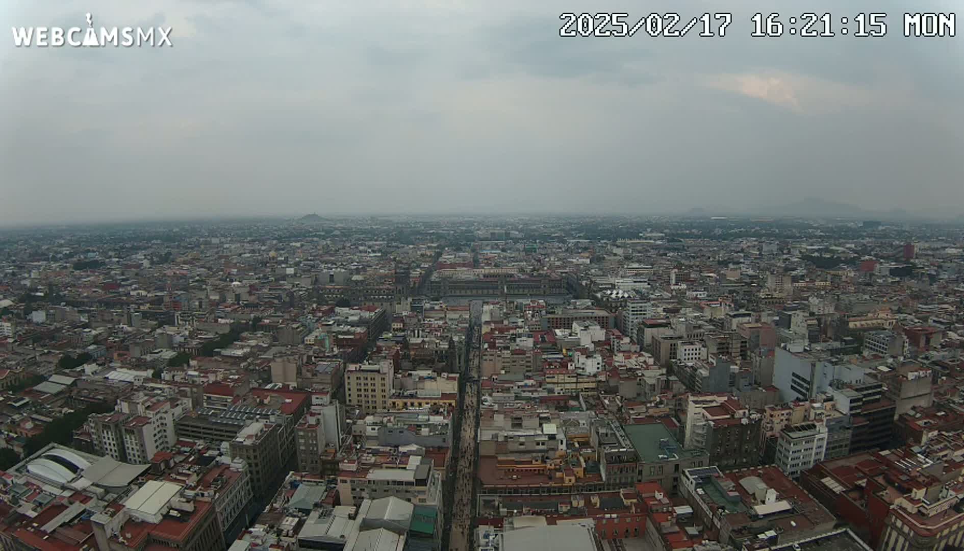 A high-angle, long shot of a sprawling city under an overcast sky shows a dense collection of buildings extending to the horizon.