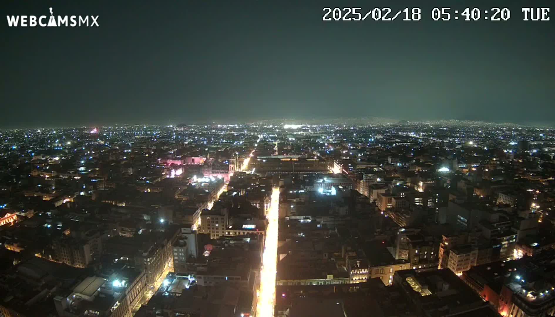 A nighttime, high-angle view shows a sprawling city illuminated by countless lights, with a brightly lit street running centrally through the scene.