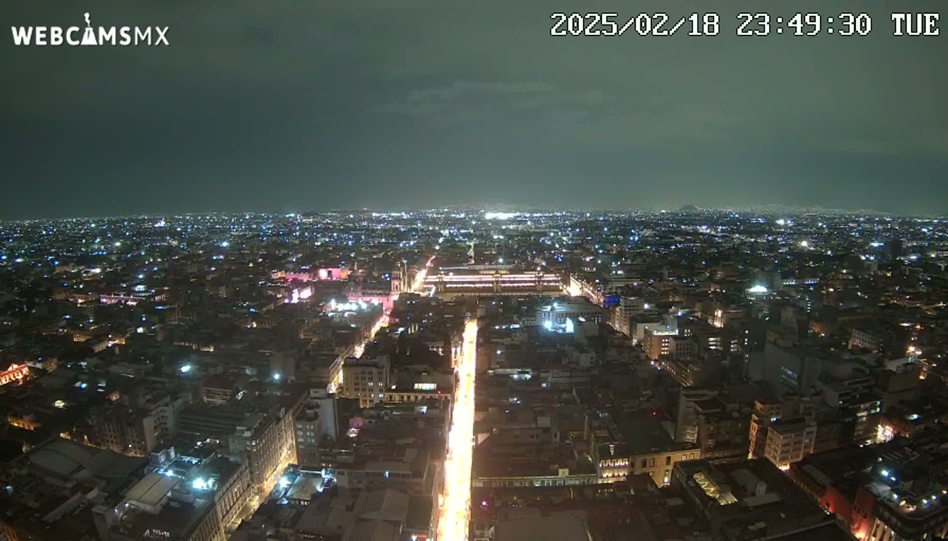 A nighttime aerial view of a sprawling city with a brightly lit street running through its center under a dark, cloudy sky.