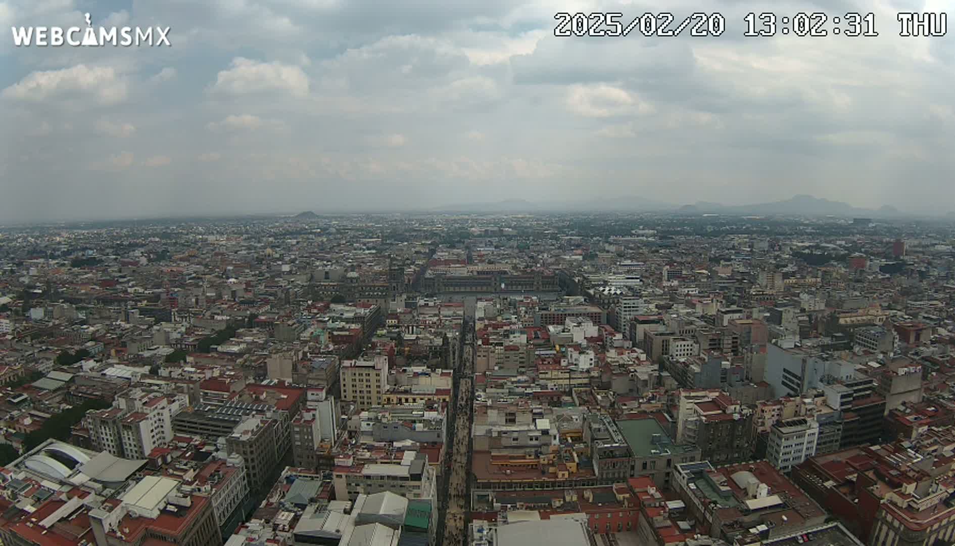 An expansive, high-angle view of a dense city under a cloudy sky shows a wide cityscape stretching to distant mountains.