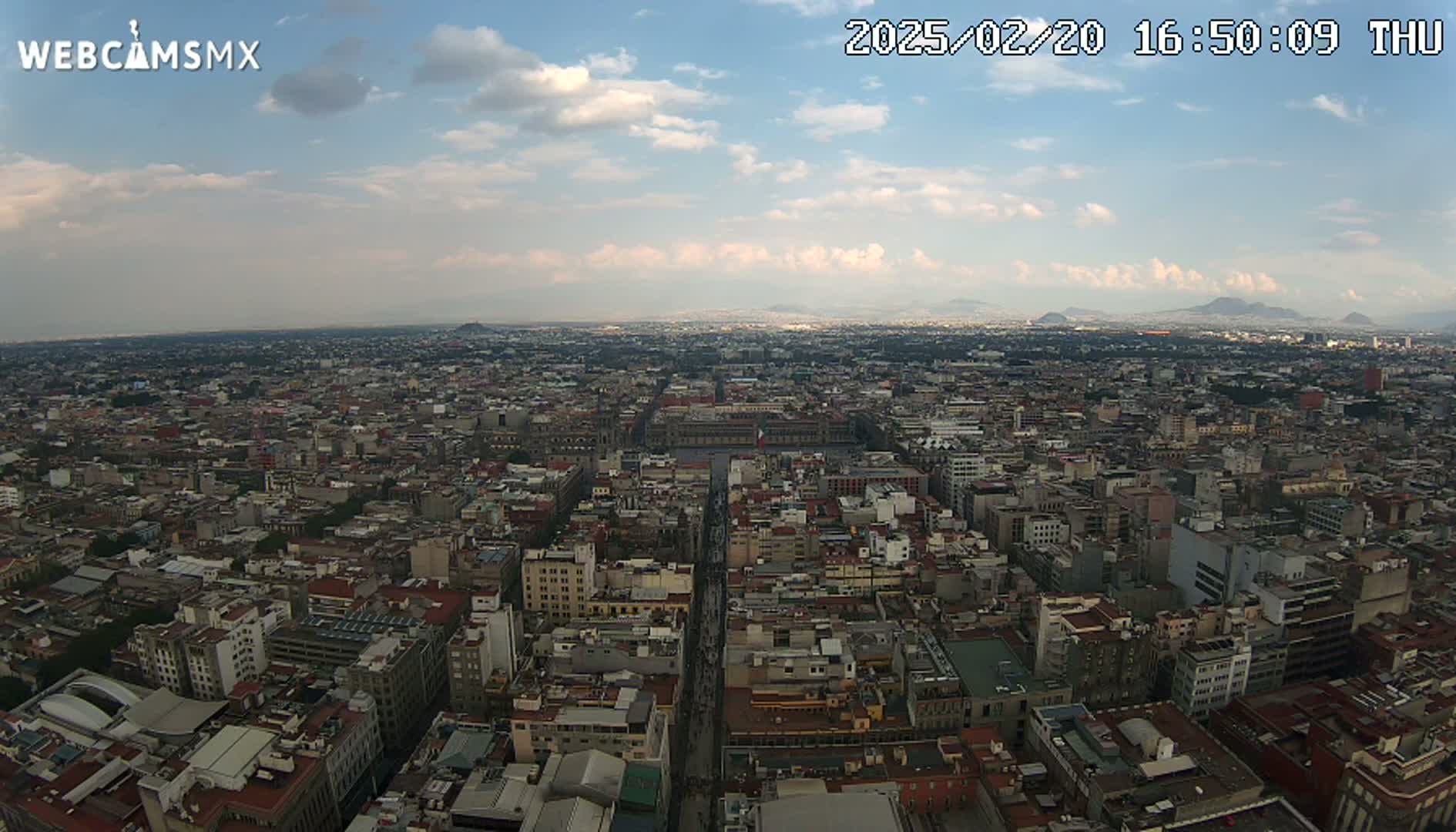 A high-angle, wide view of a sprawling city on a partly cloudy day shows a large plaza in the middle of densely packed buildings extending to the horizon, with distant mountains visible.