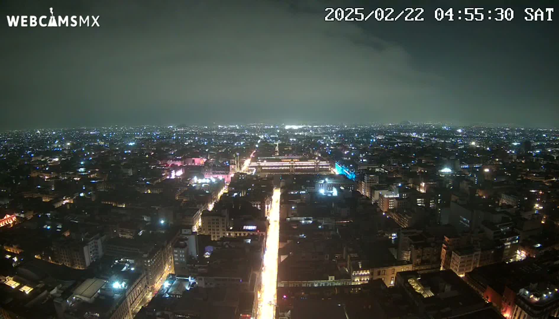 A nighttime, aerial view of a sprawling city with a brightly lit central avenue extending towards the horizon under a dark, cloudy sky.