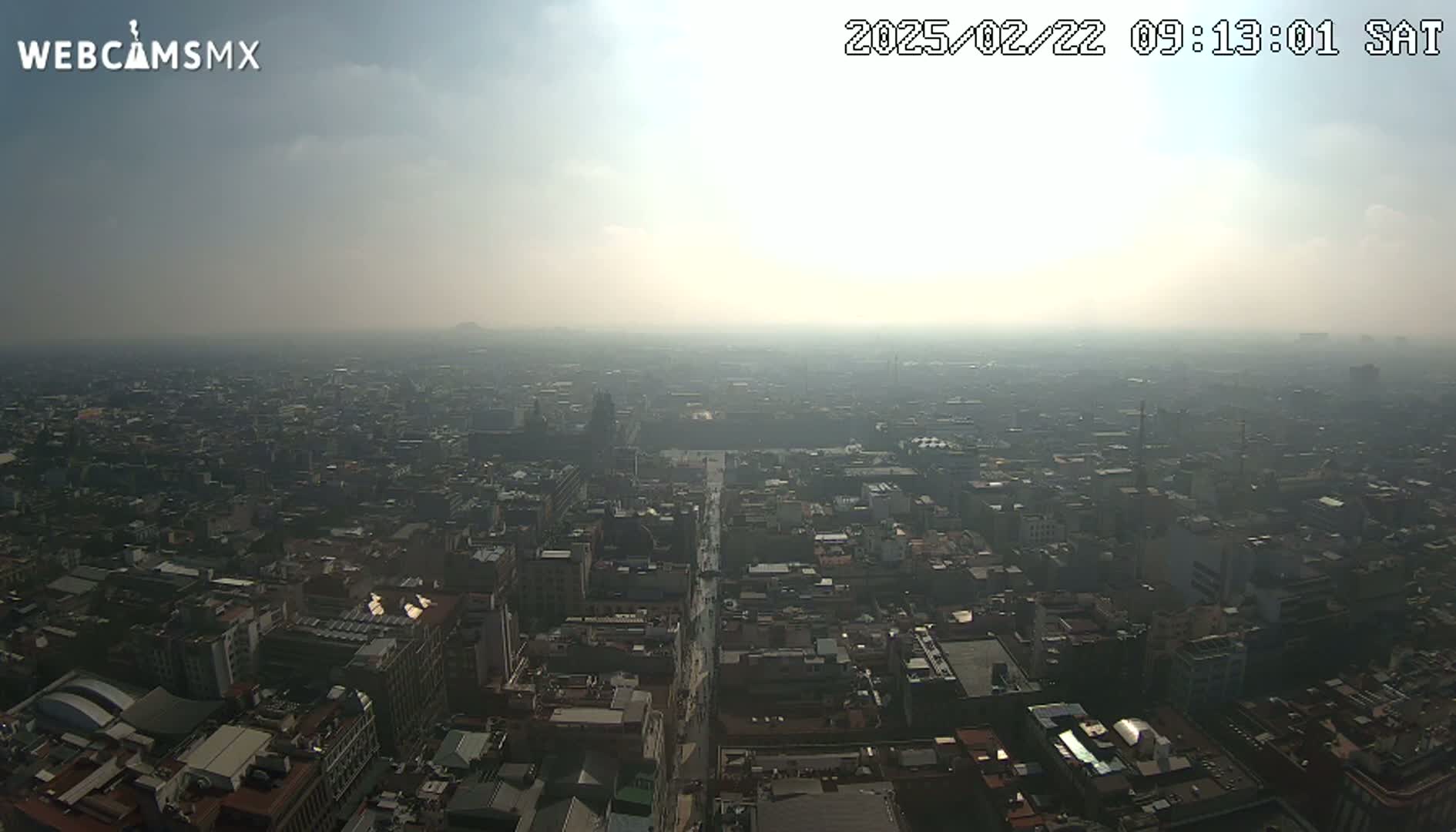A hazy, sunny, high-angle view of a sprawling city with a long, straight road cutting through its center.