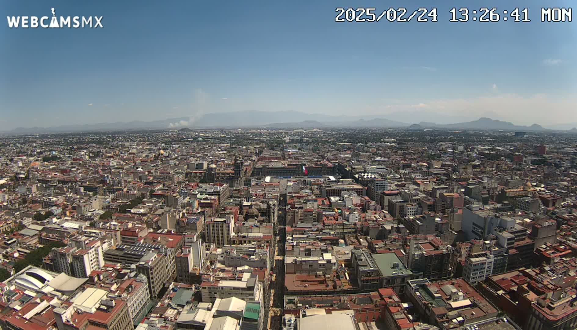A high-angle, long shot on a clear day shows a sprawling city with low-rise buildings extending to distant mountains under a mostly clear sky.