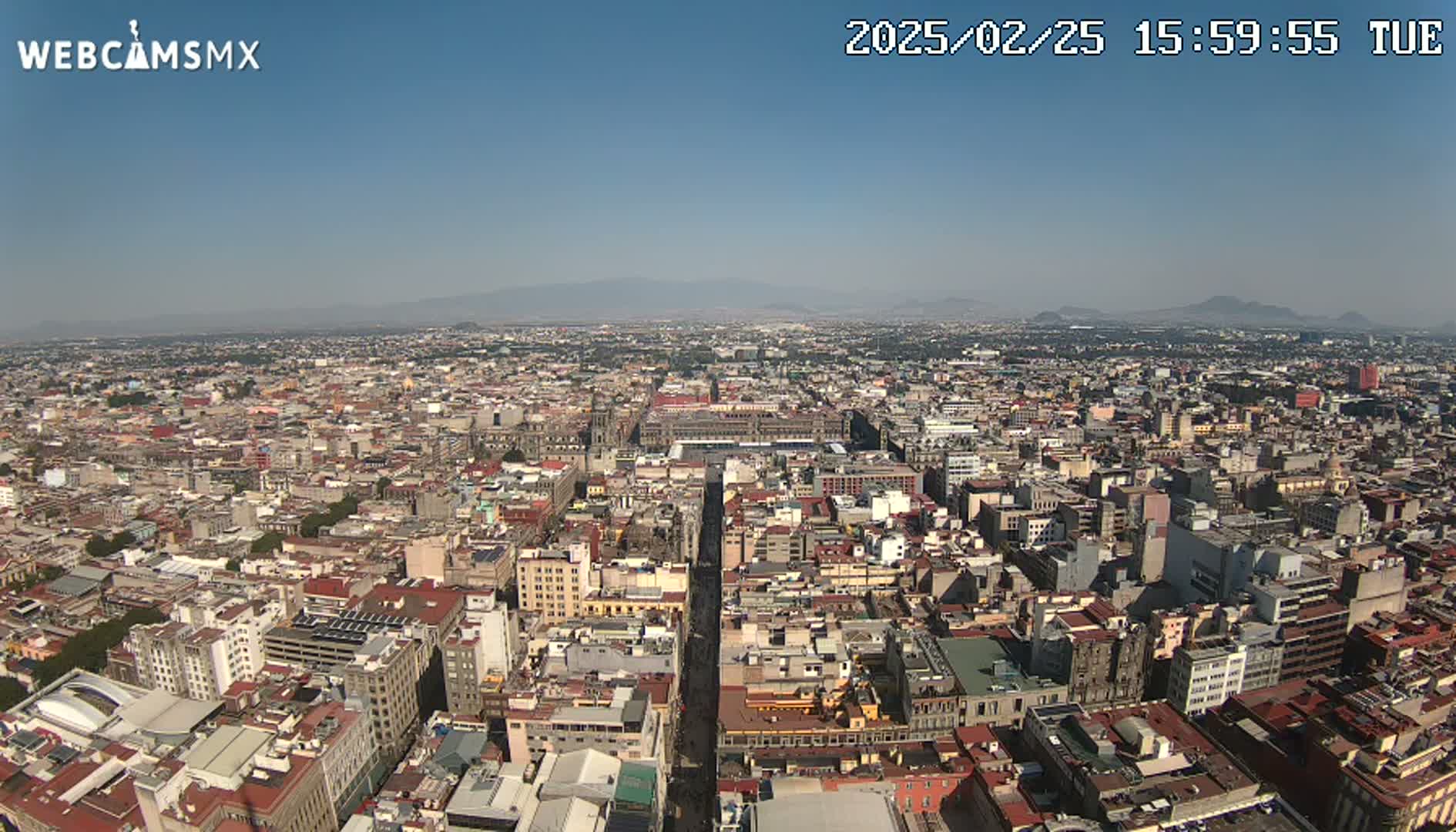 A high-angle, long shot of a sprawling city on a clear, sunny day, with distant mountains visible on the horizon.