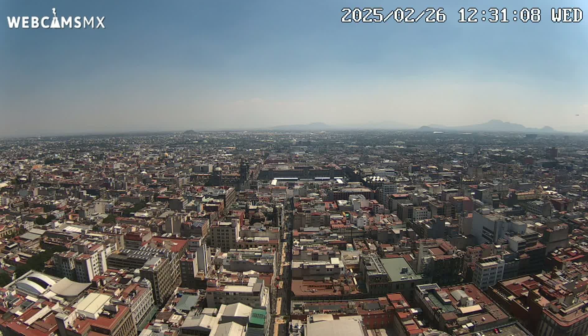 A high-angle, long shot on a clear, sunny day shows a sprawling cityscape extending to distant mountains.