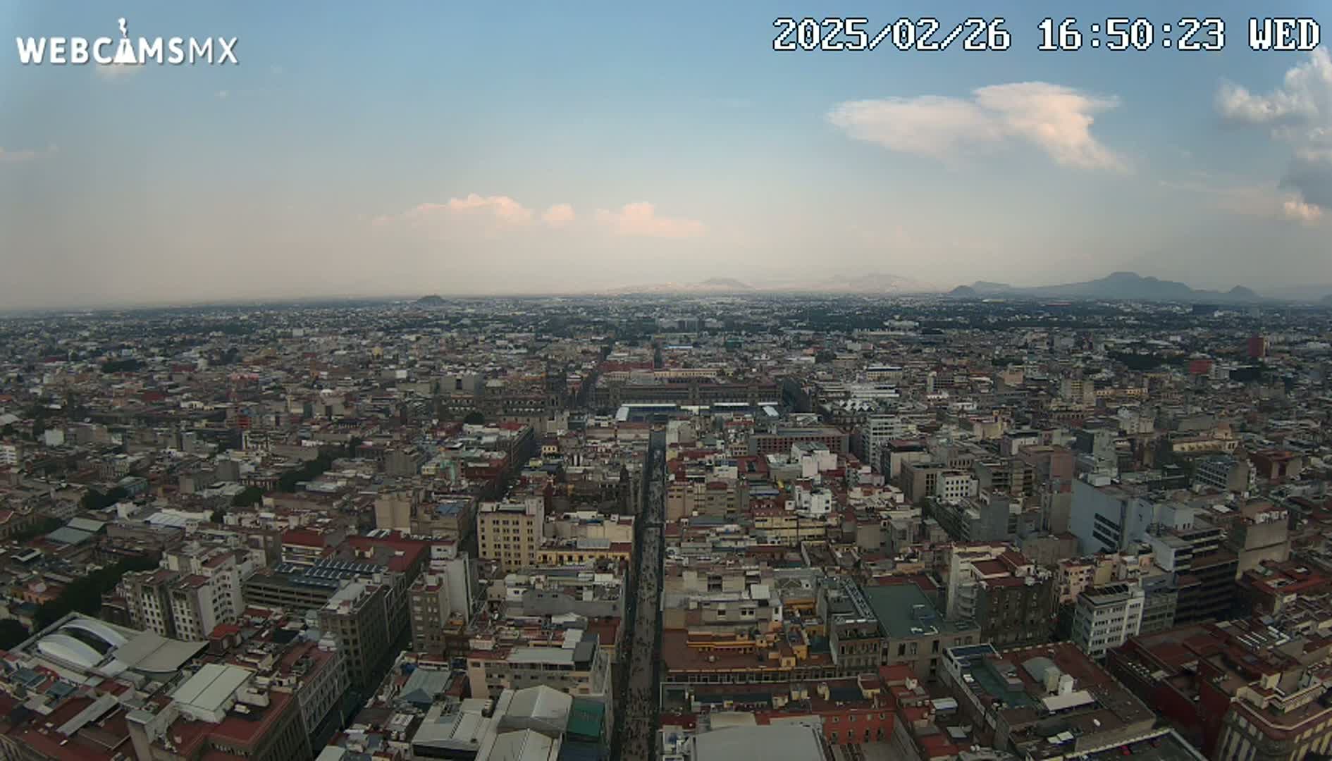 A high-angle, long shot of a sprawling city under a partly cloudy sky shows a vast urban landscape extending to distant mountains.