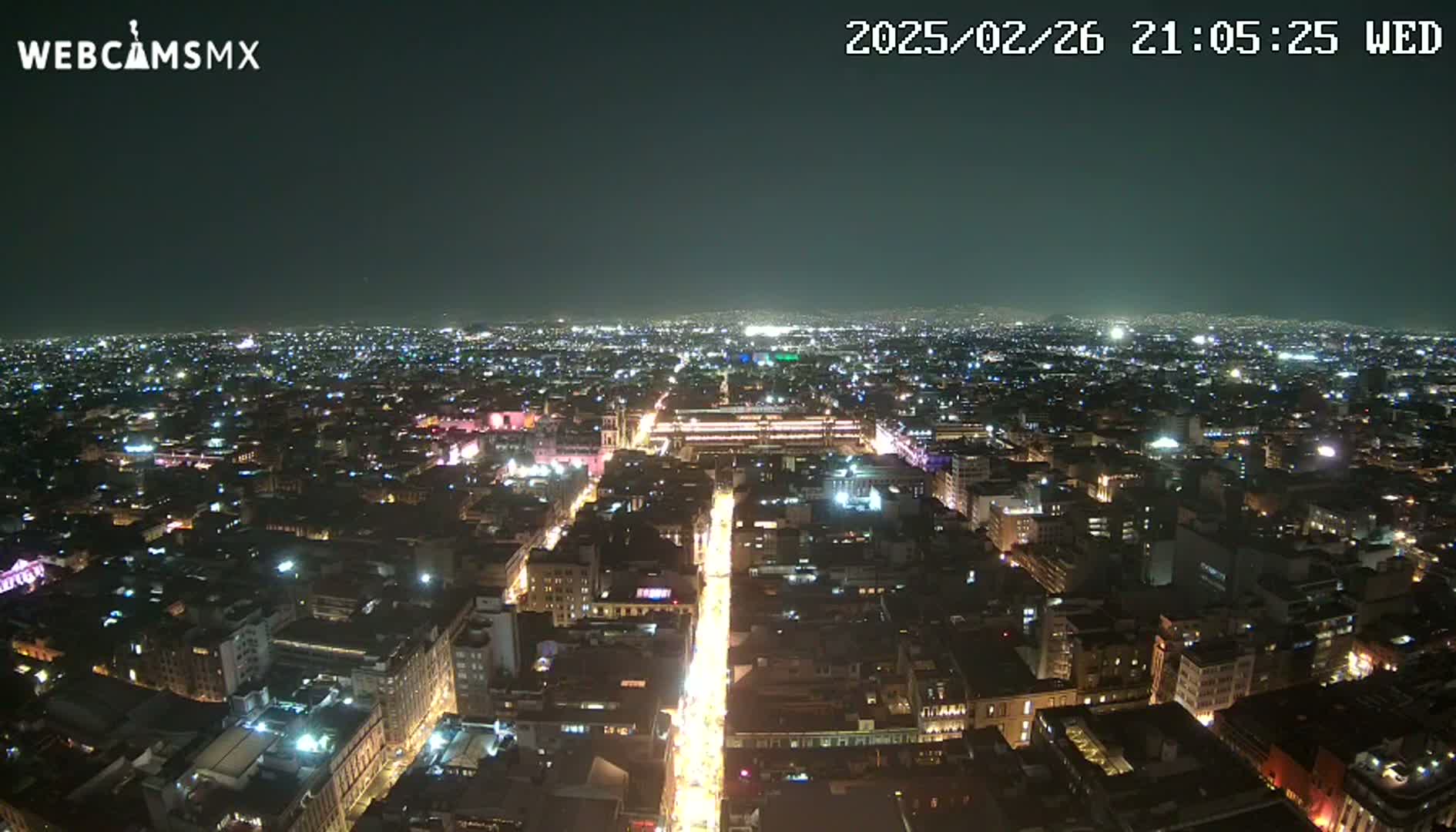 A nighttime aerial view shows a sprawling city illuminated by countless lights, with a brightly lit street running through the center.