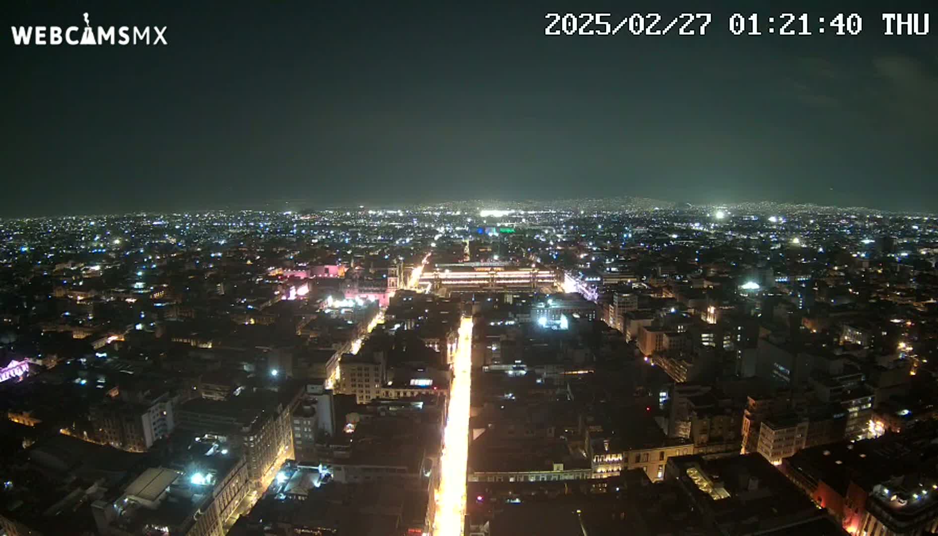 A brightly lit city at night is viewed from above, showing a long, illuminated street extending from the center of the image toward the viewer.