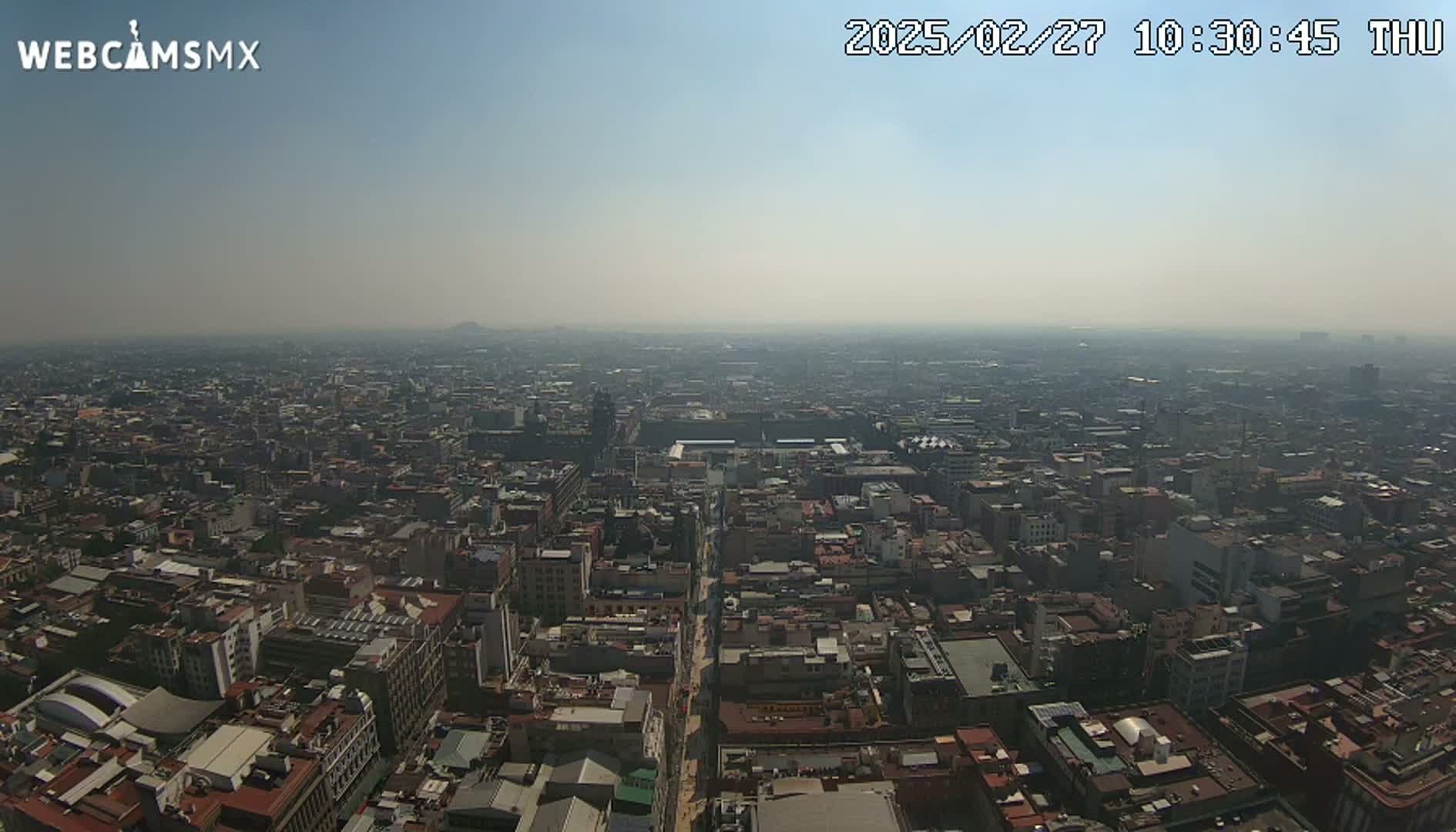 A hazy, high-angle view shows a vast expanse of a city with densely packed buildings stretching to the horizon under a clear, light-blue sky.