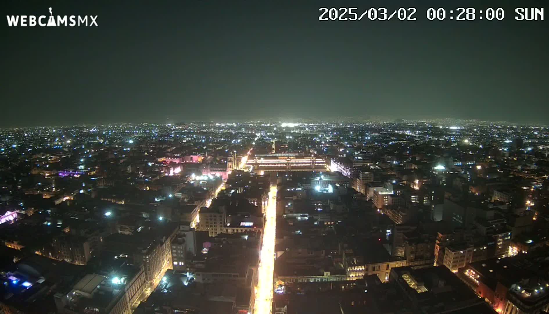 A brightly lit city street at night extends from a high vantage point into a vast expanse of dimly lit buildings under a clear, dark sky.