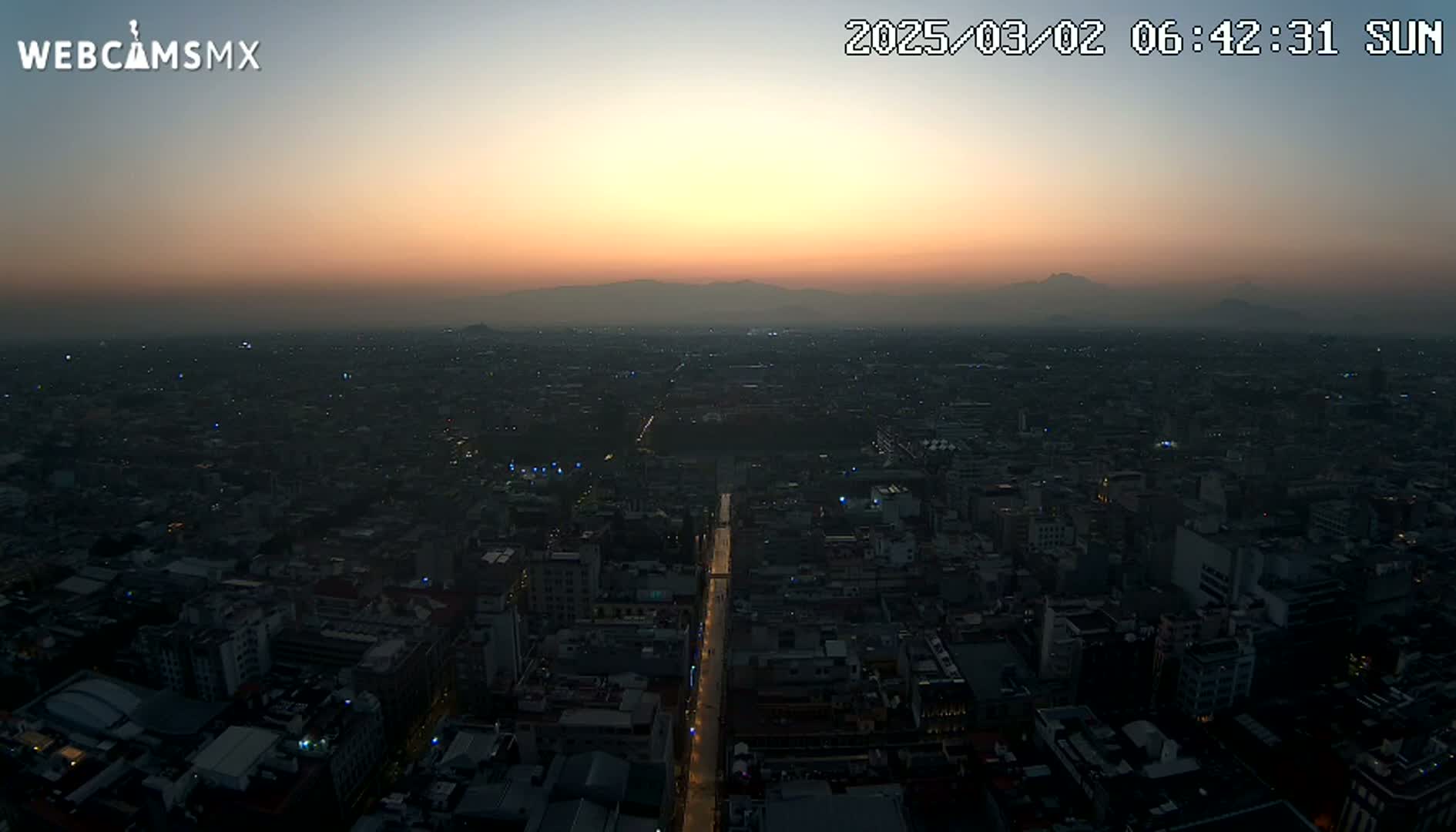 A hazy sunset illuminates a sprawling city at twilight, with distant mountains visible on the horizon.