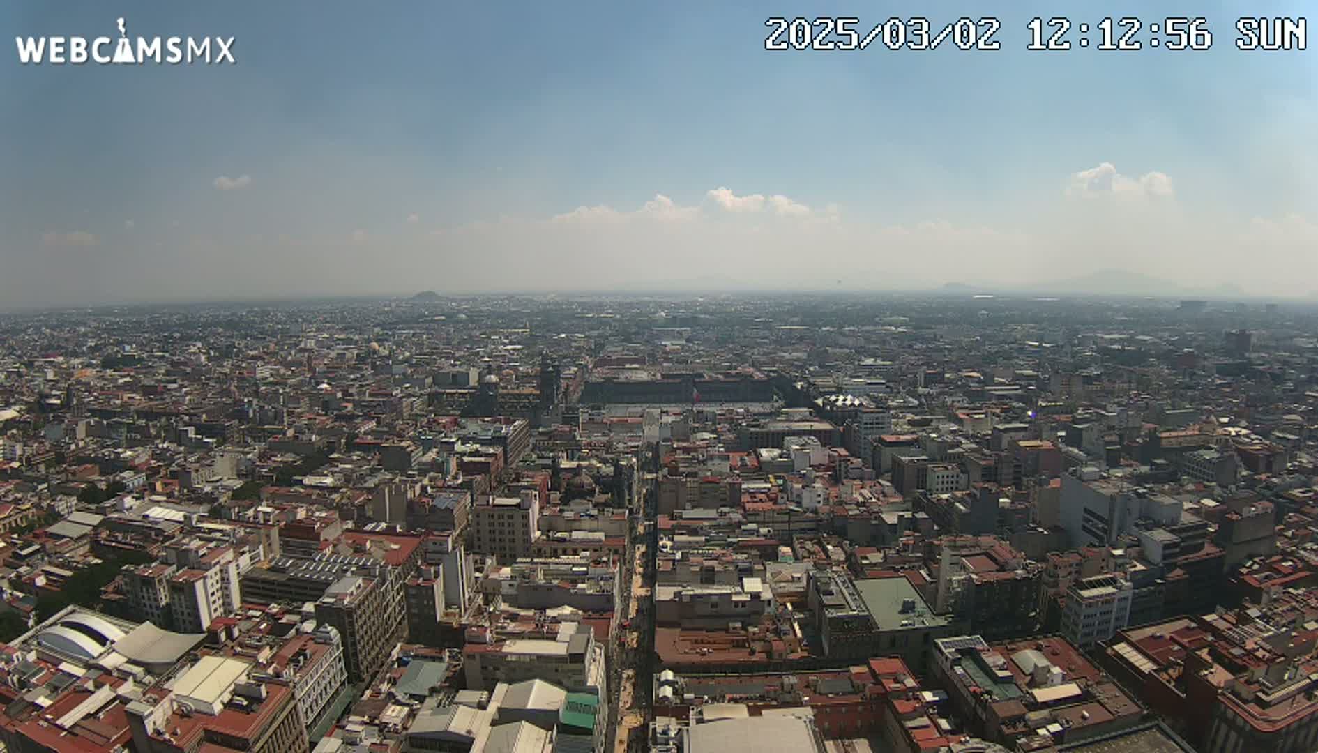 A high-angle, long shot shows a sprawling cityscape under a mostly clear, hazy sky with a few scattered clouds.