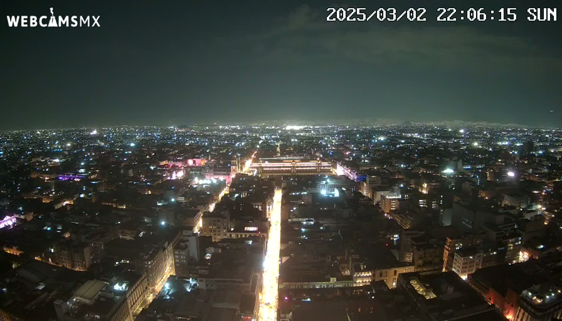 A nighttime aerial view of a city shows brightly lit streets and buildings under a mostly clear sky.