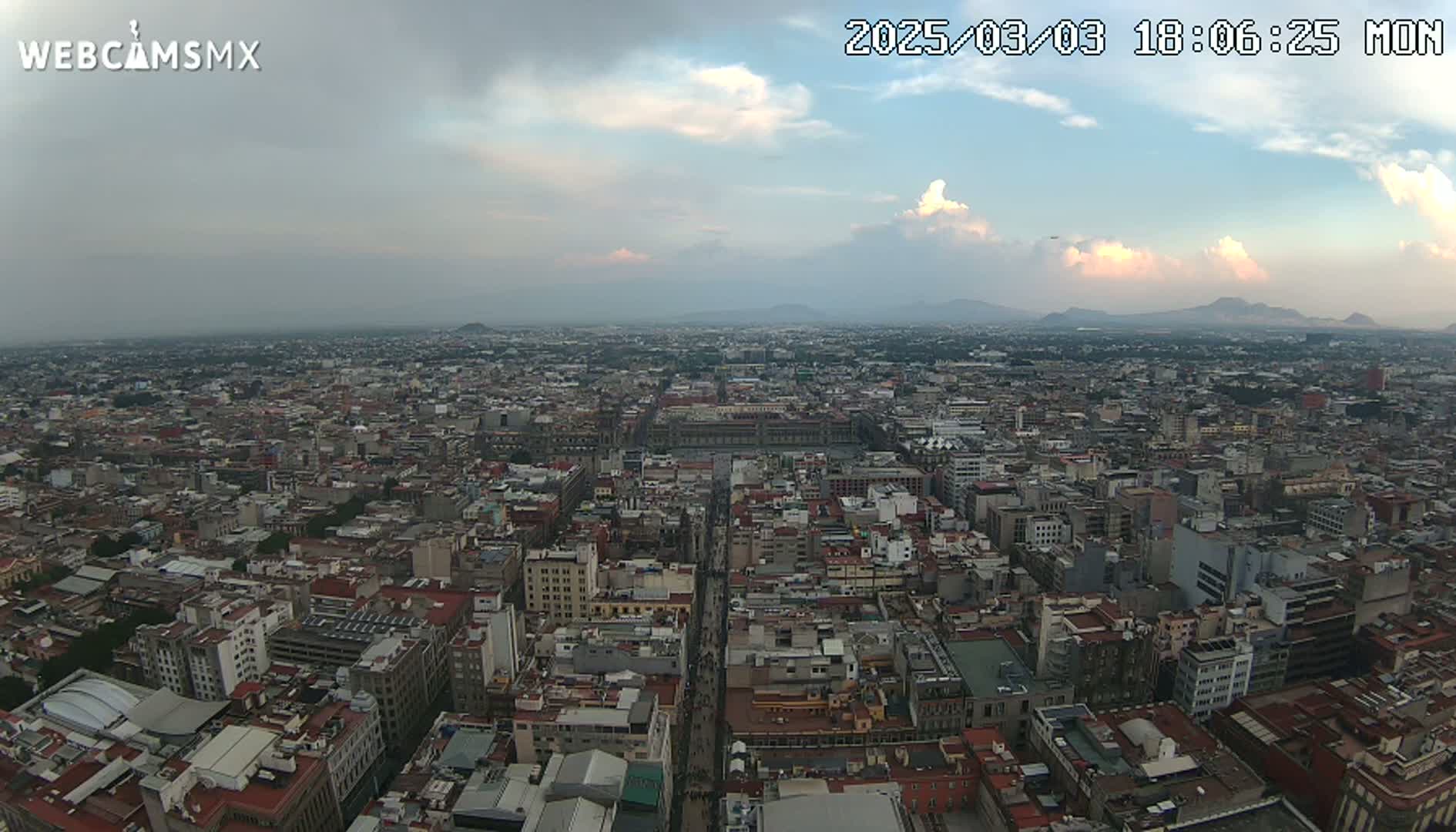 A high-angle, long shot of a sprawling city under a partly cloudy sky, with mountains visible in the distance.