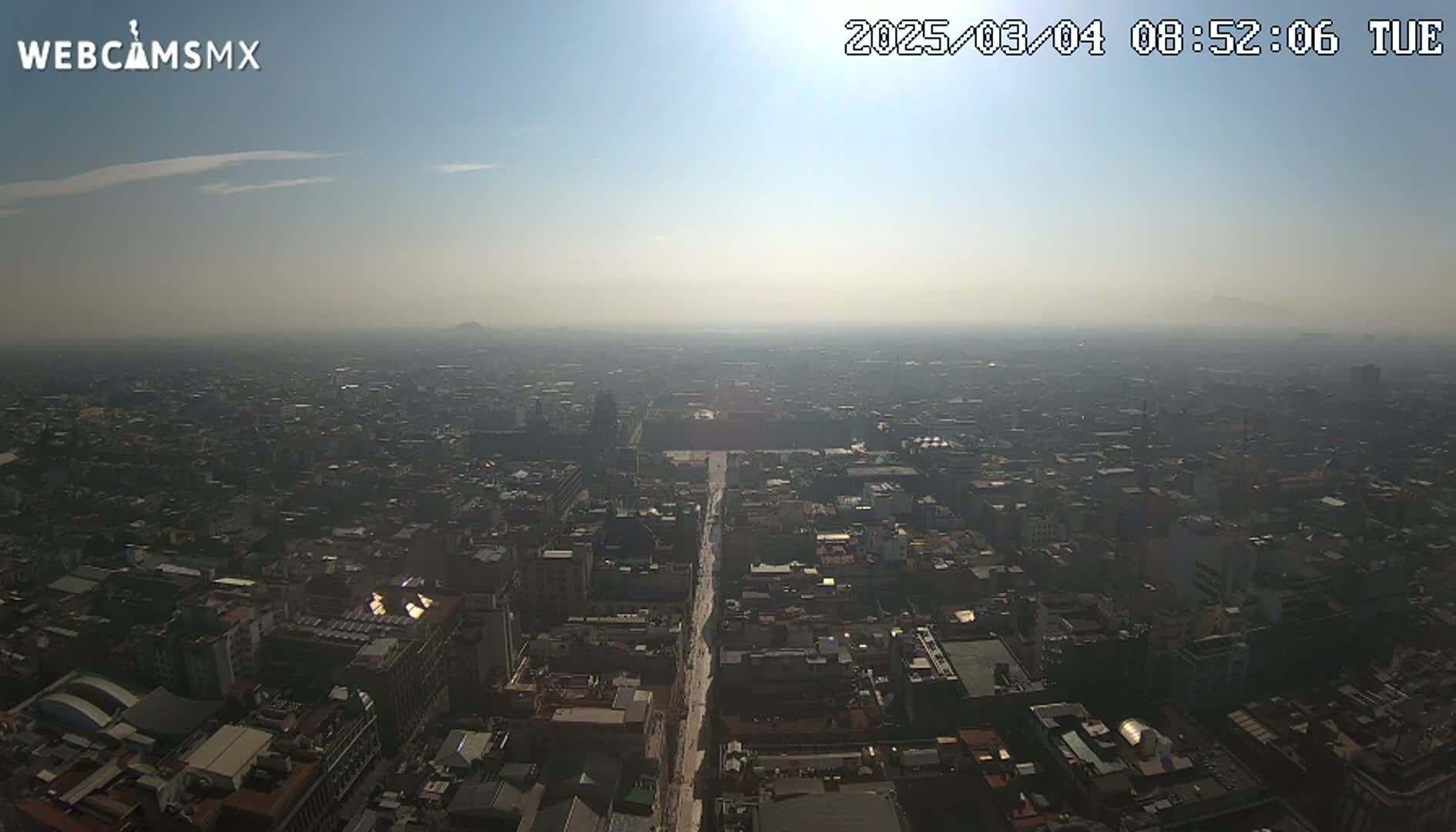 A hazy, sunny day reveals a sprawling cityscape viewed from a high vantage point, with a long, straight road visible cutting through the dense buildings.