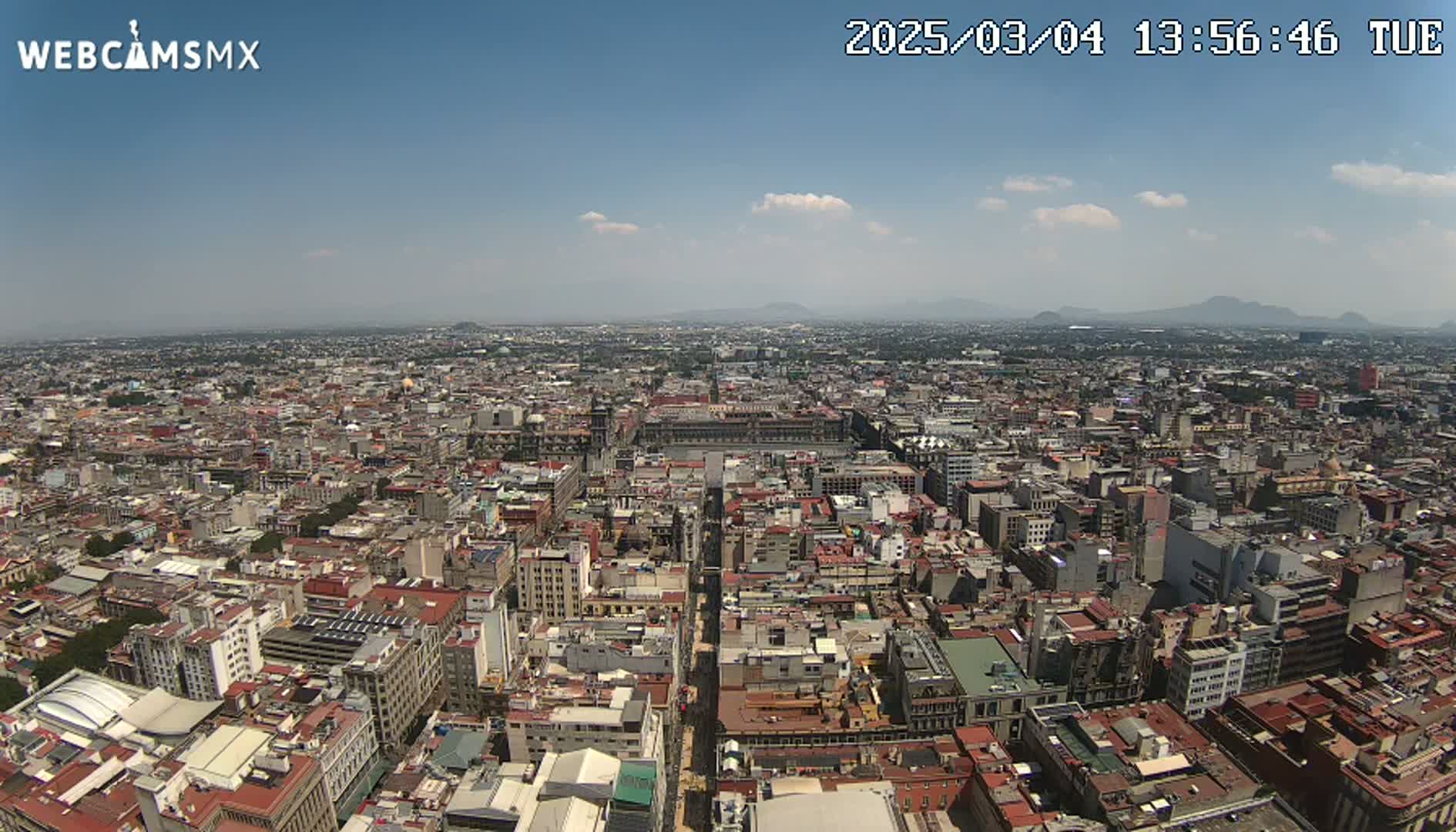 A high-angle, long shot of a sprawling city under a mostly clear, sunny sky with a few small clouds and distant mountains in the background.