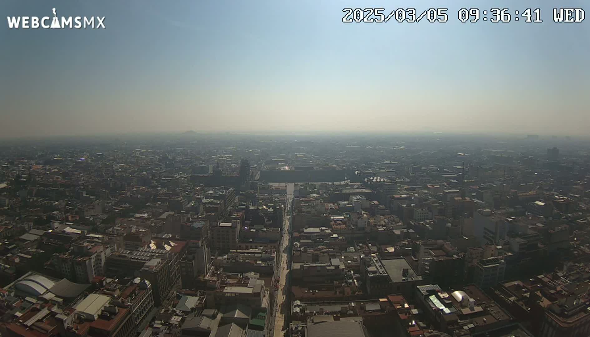 A hazy, sunny aerial view of a vast city with a long street cutting through its center.
