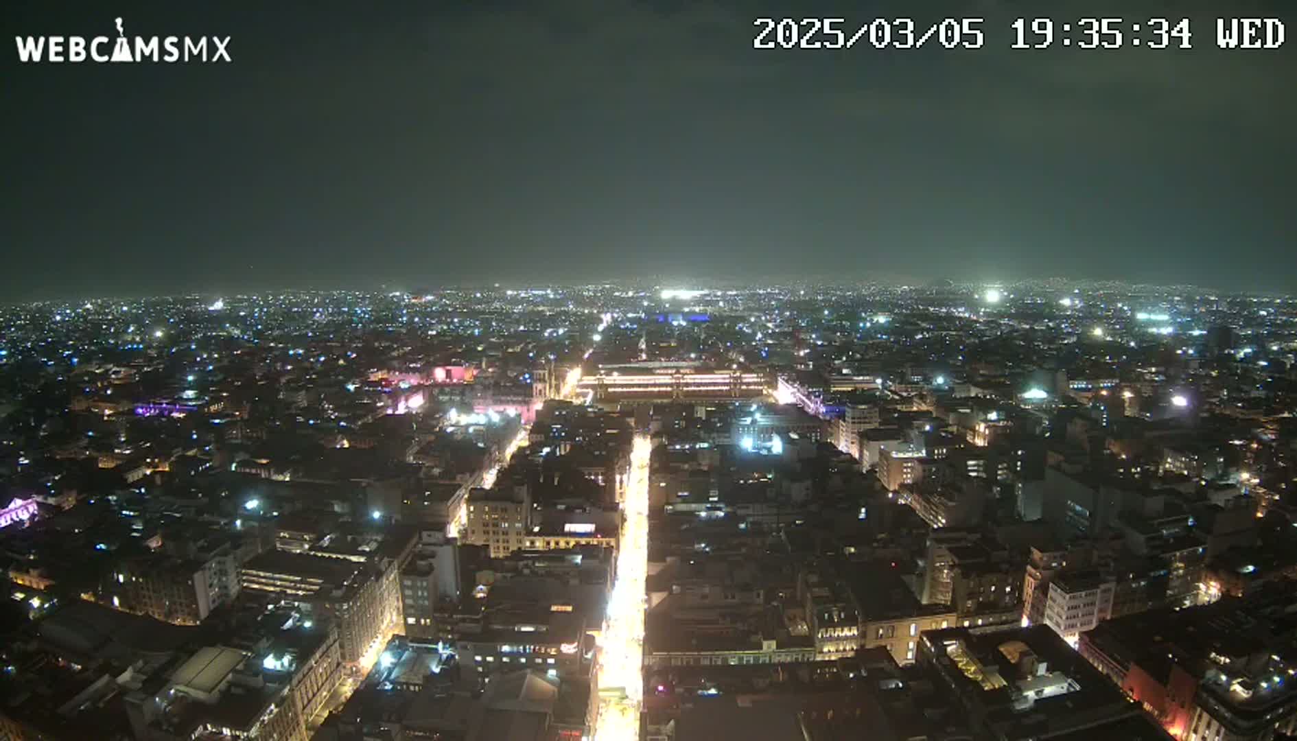 A nighttime, high-angle view shows a sprawling city illuminated by countless lights, with a brightly lit street cutting through the urban landscape.