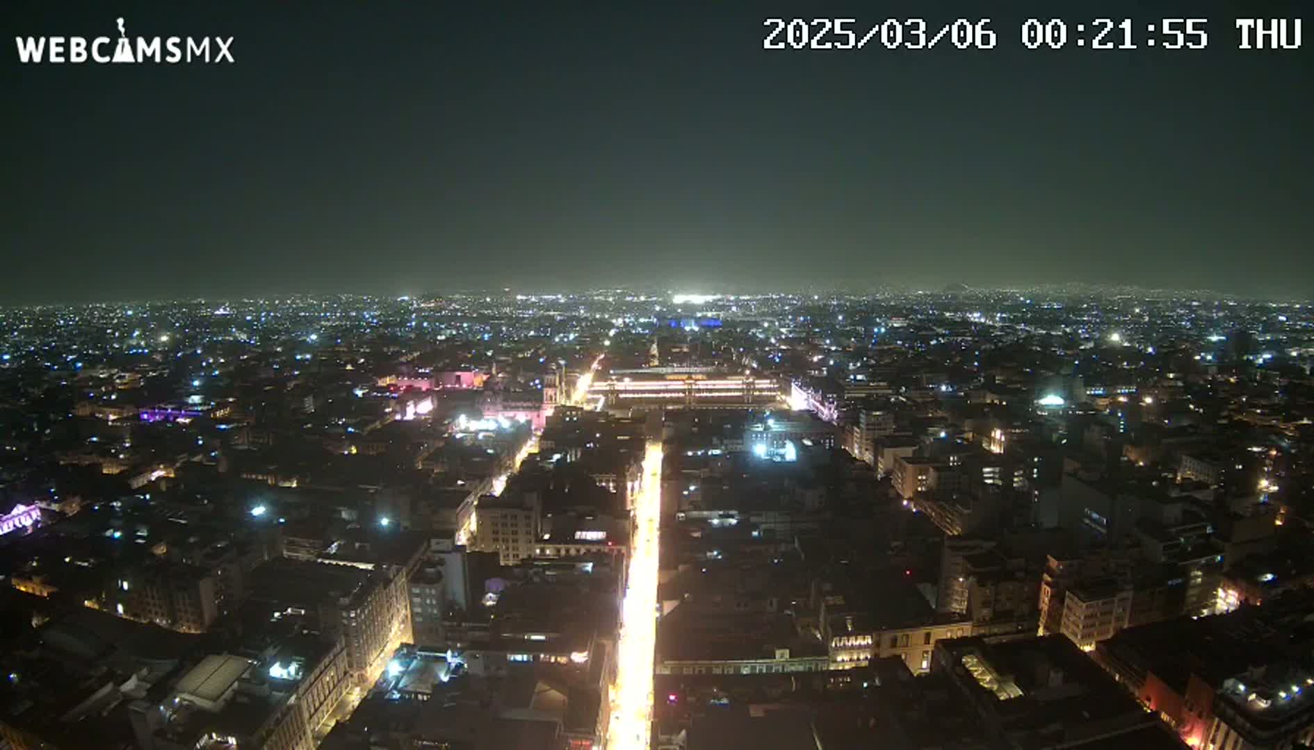 A nighttime, high-angle view of a sprawling city illuminated by countless lights, with a brightly lit avenue stretching from the foreground into the distance.