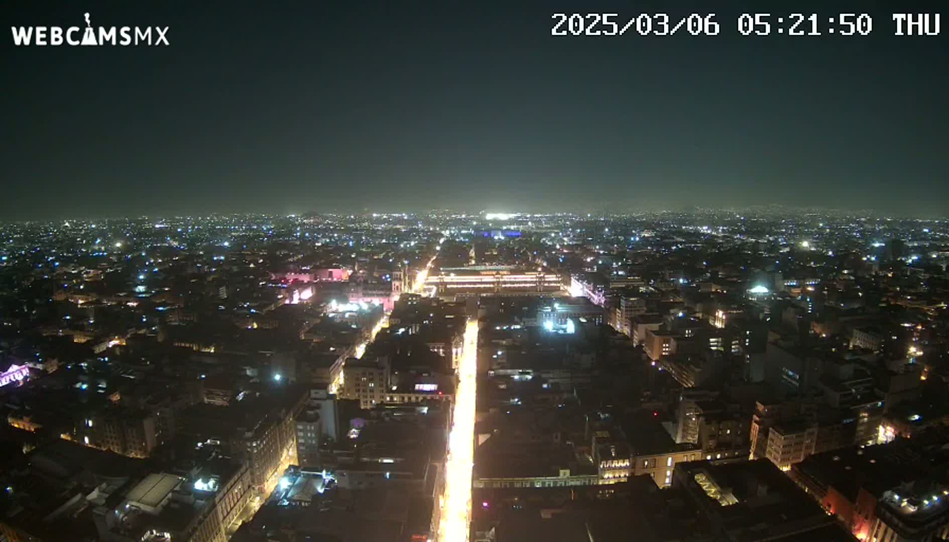 A brightly lit city street at night stretches into the distance, surrounded by a sprawling cityscape under a clear, dark sky.