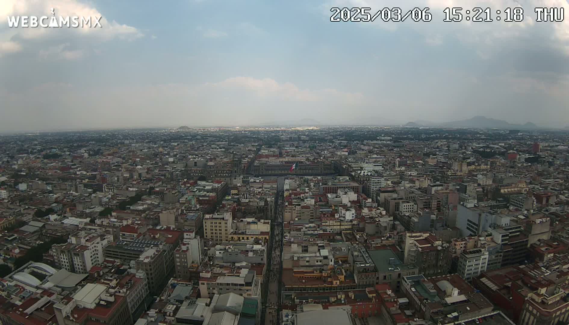 An aerial view of a sprawling city under a cloudy sky shows a large plaza in the center with a street extending from it, surrounded by dense urban development.