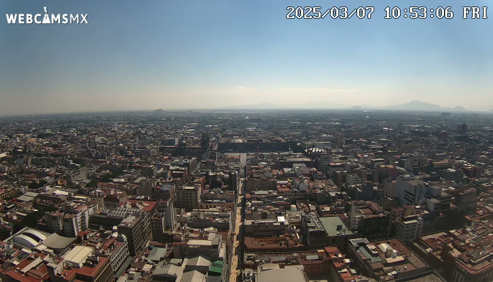 A high-angle, long shot on a clear, sunny day shows a sprawling cityscape extending to distant mountains under a mostly clear sky.