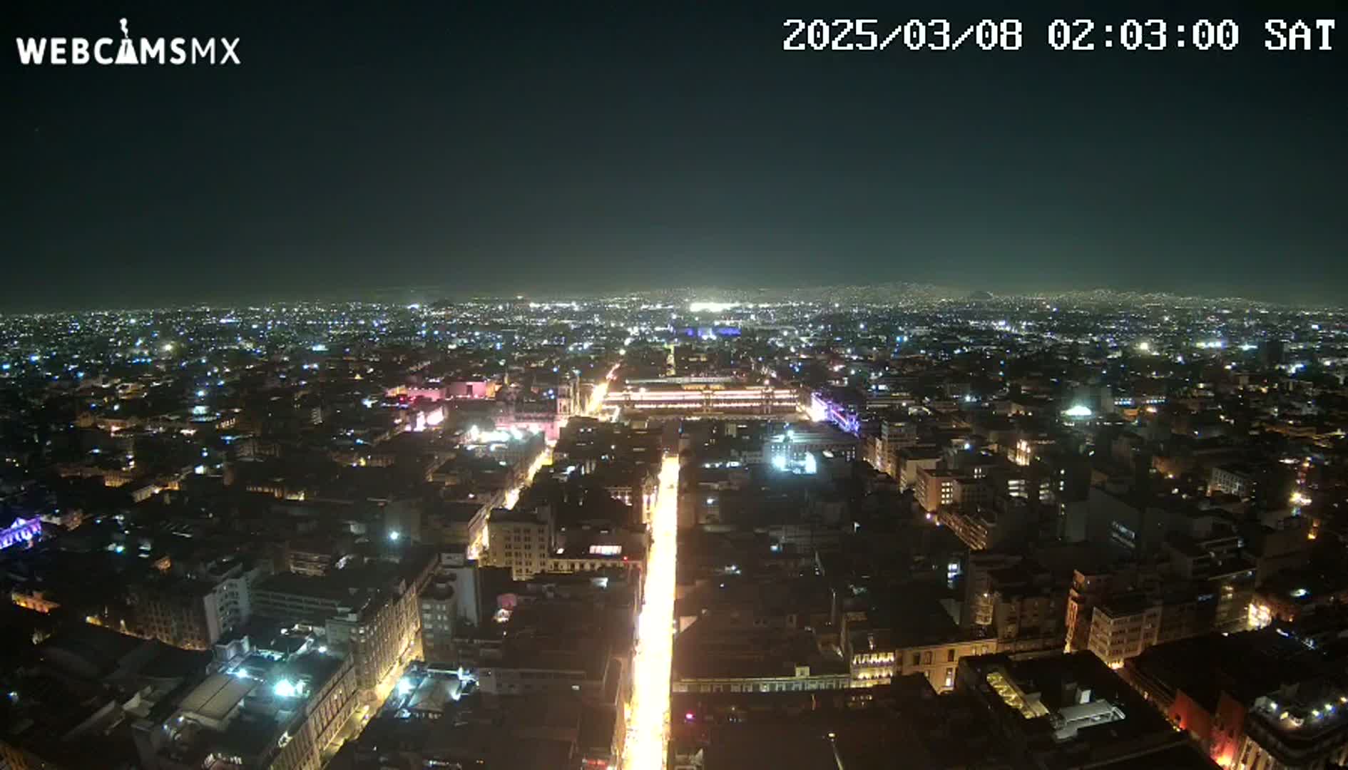 A nighttime, high-angle view shows a brightly lit city street extending from the foreground toward the distant, softly illuminated cityscape under a dark, clear sky.