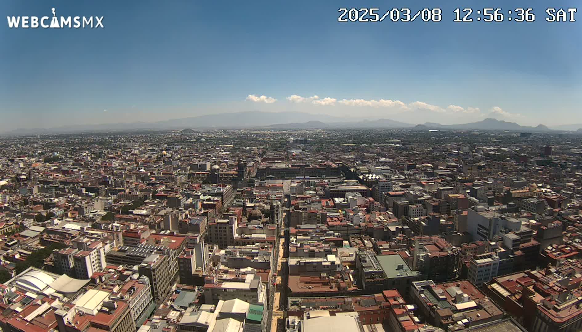 A high-angle, long shot on a clear, sunny day shows a vast cityscape sprawling to distant mountains under a mostly clear blue sky.
