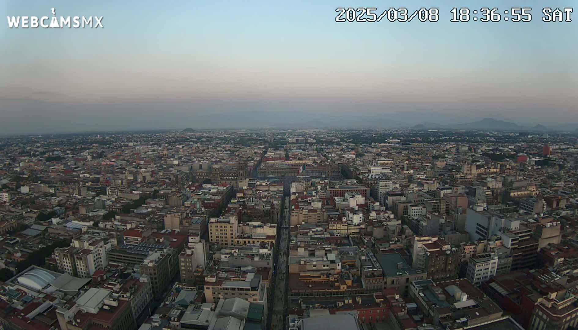 A hazy dusk settles over a sprawling city, viewed from a high vantage point, revealing a dense network of buildings extending to distant mountains.