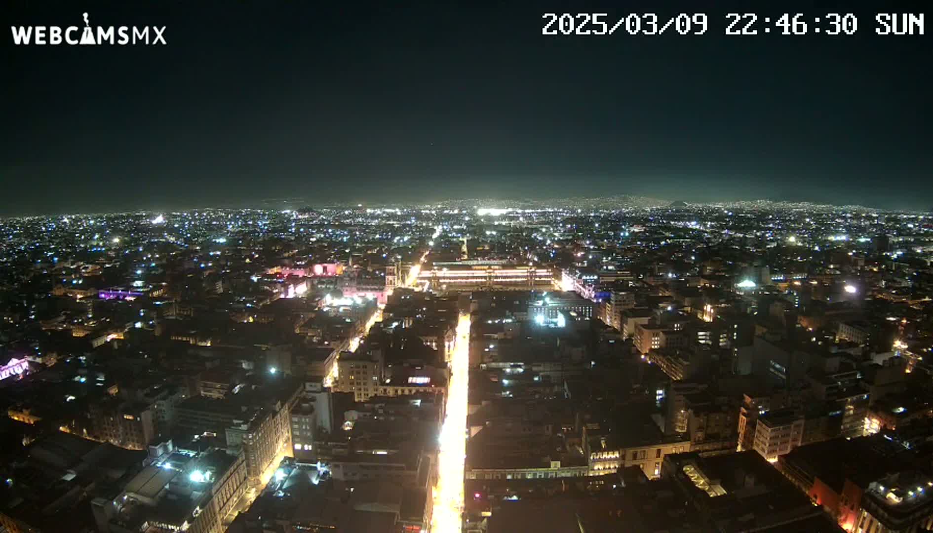 A nighttime aerial view shows a sprawling city illuminated by countless lights, with a brightly lit avenue extending from the foreground into the distance under a clear, dark sky.