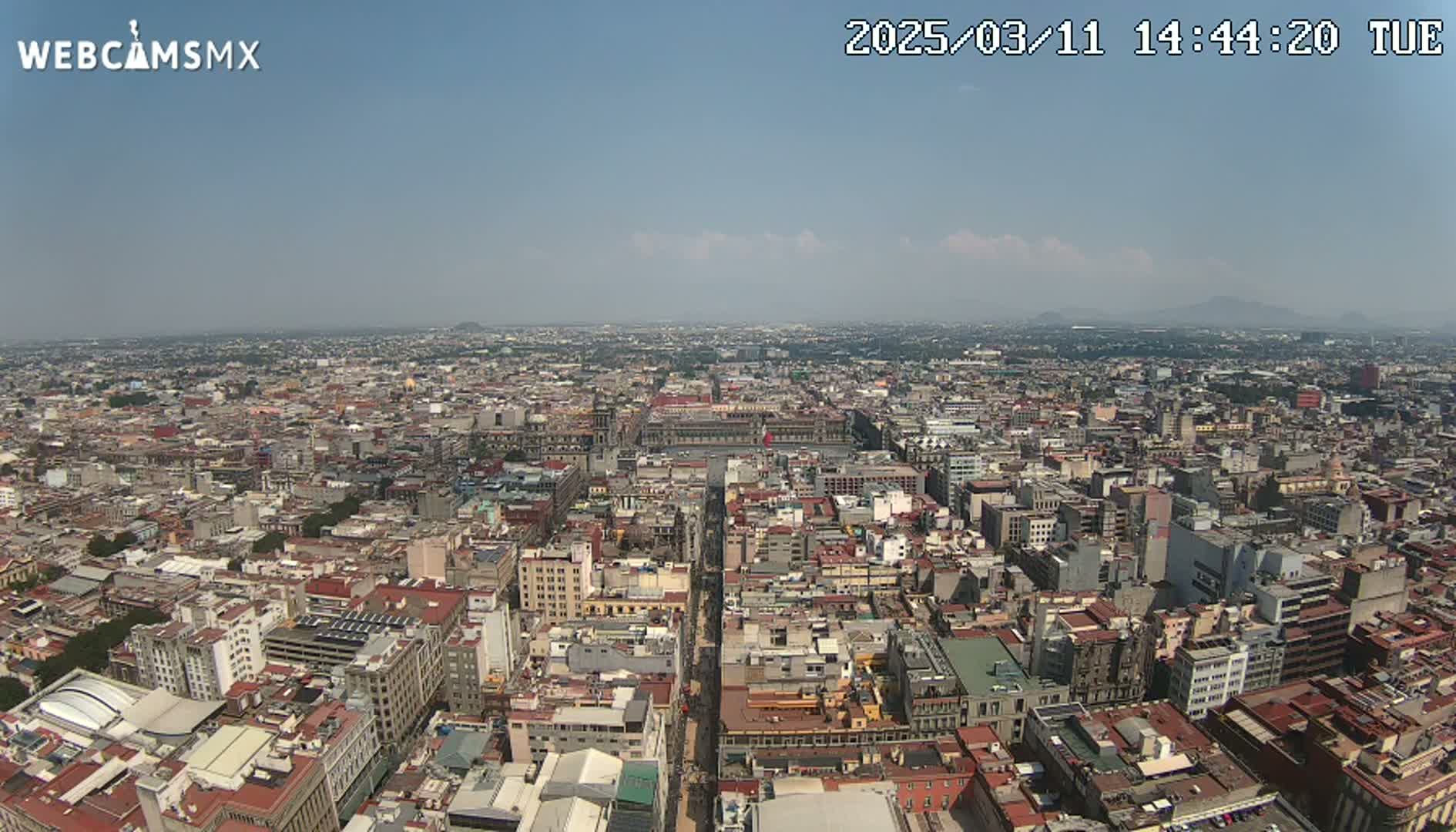 A high-angle, long shot on a clear, sunny day shows a sprawling city with many closely packed buildings, extending to distant mountains on the horizon.