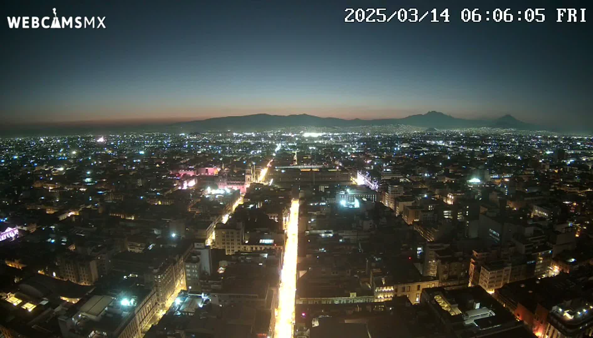 A nighttime aerial view of a city illuminated by streetlights, with distant mountains visible under a clear, dark sky.