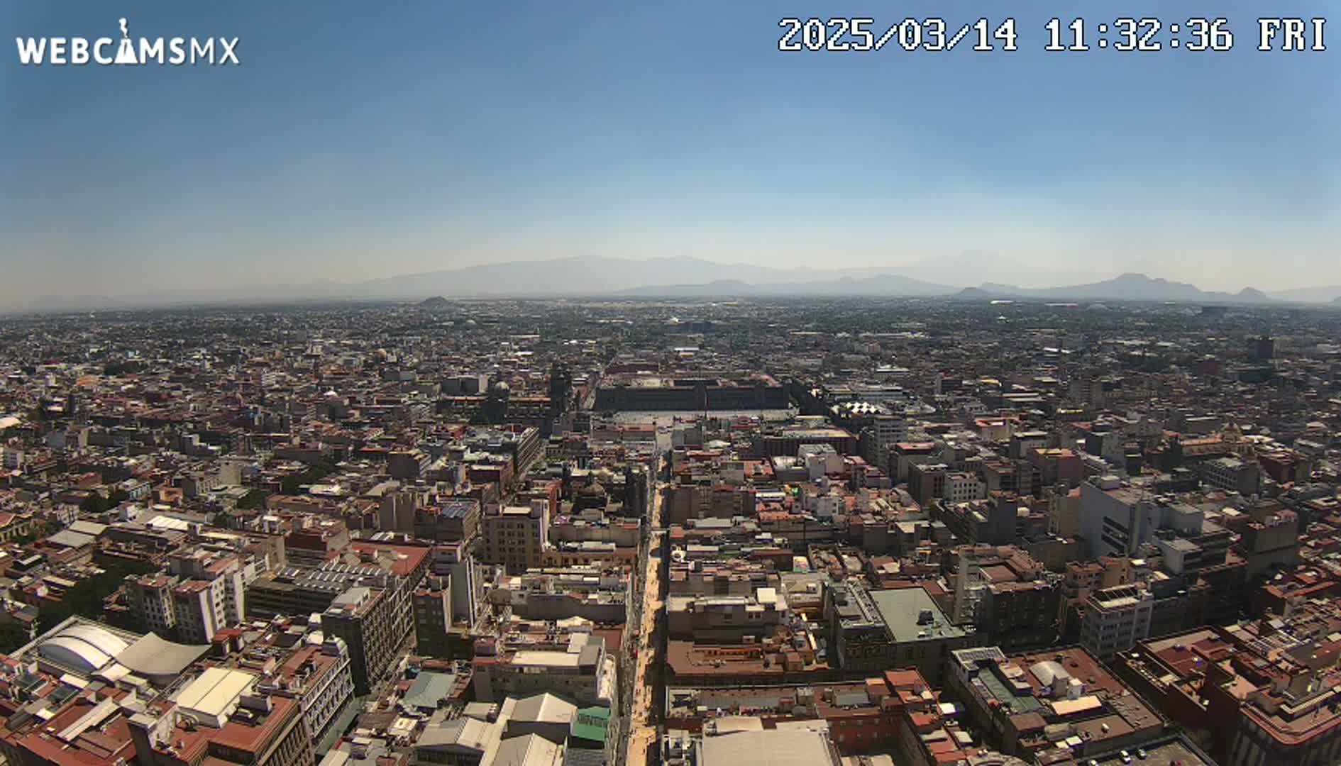 A high-angle, long shot on a clear, sunny day shows a sprawling cityscape with distant mountains under a mostly clear sky.