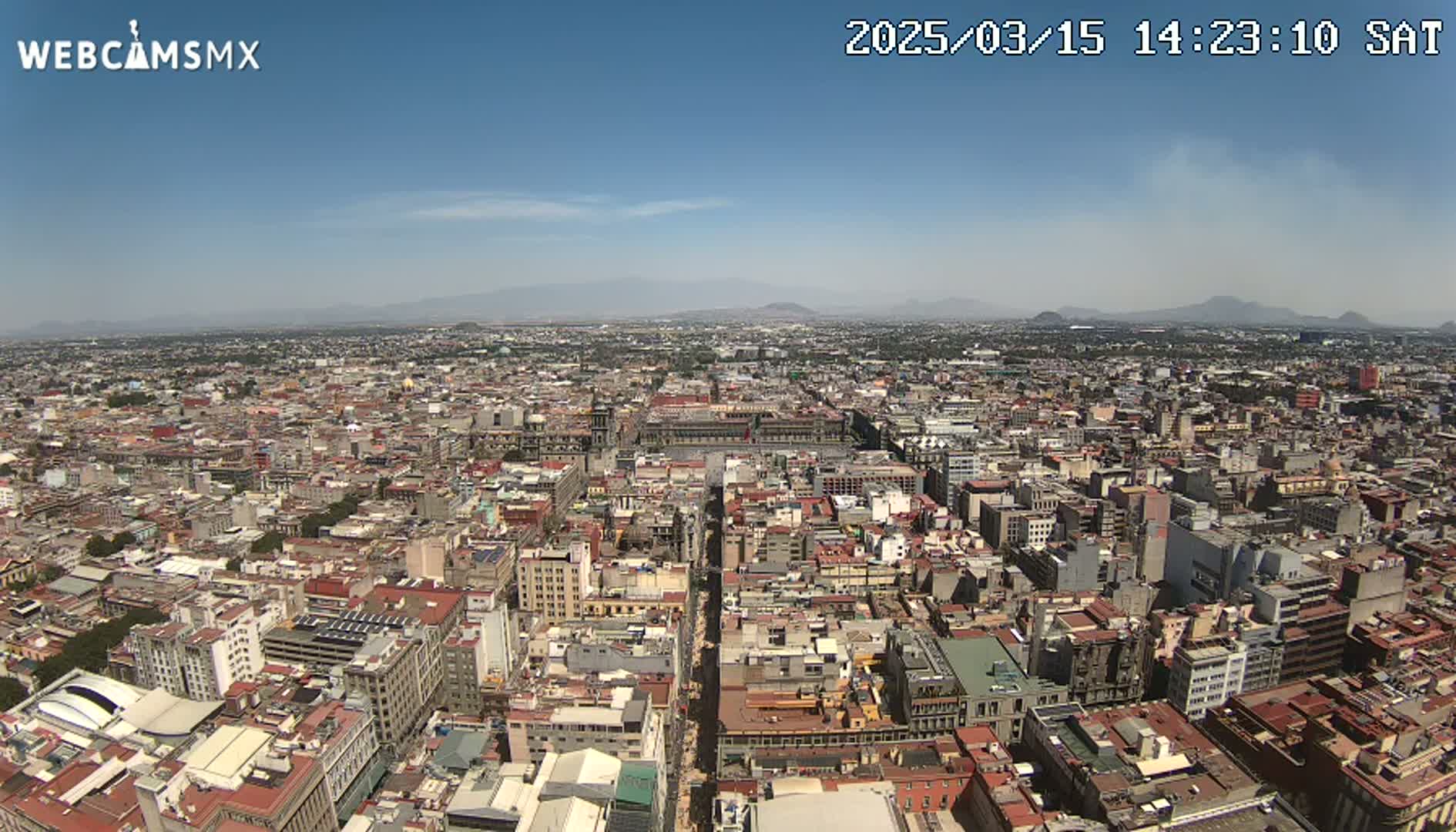 A high-angle, long shot on a clear day shows a sprawling city with low-rise buildings extending to distant mountains under a mostly clear blue sky.